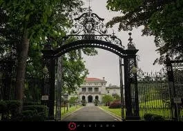 Gate leading to a large estate with a mansion in the background, surrounded by trees.