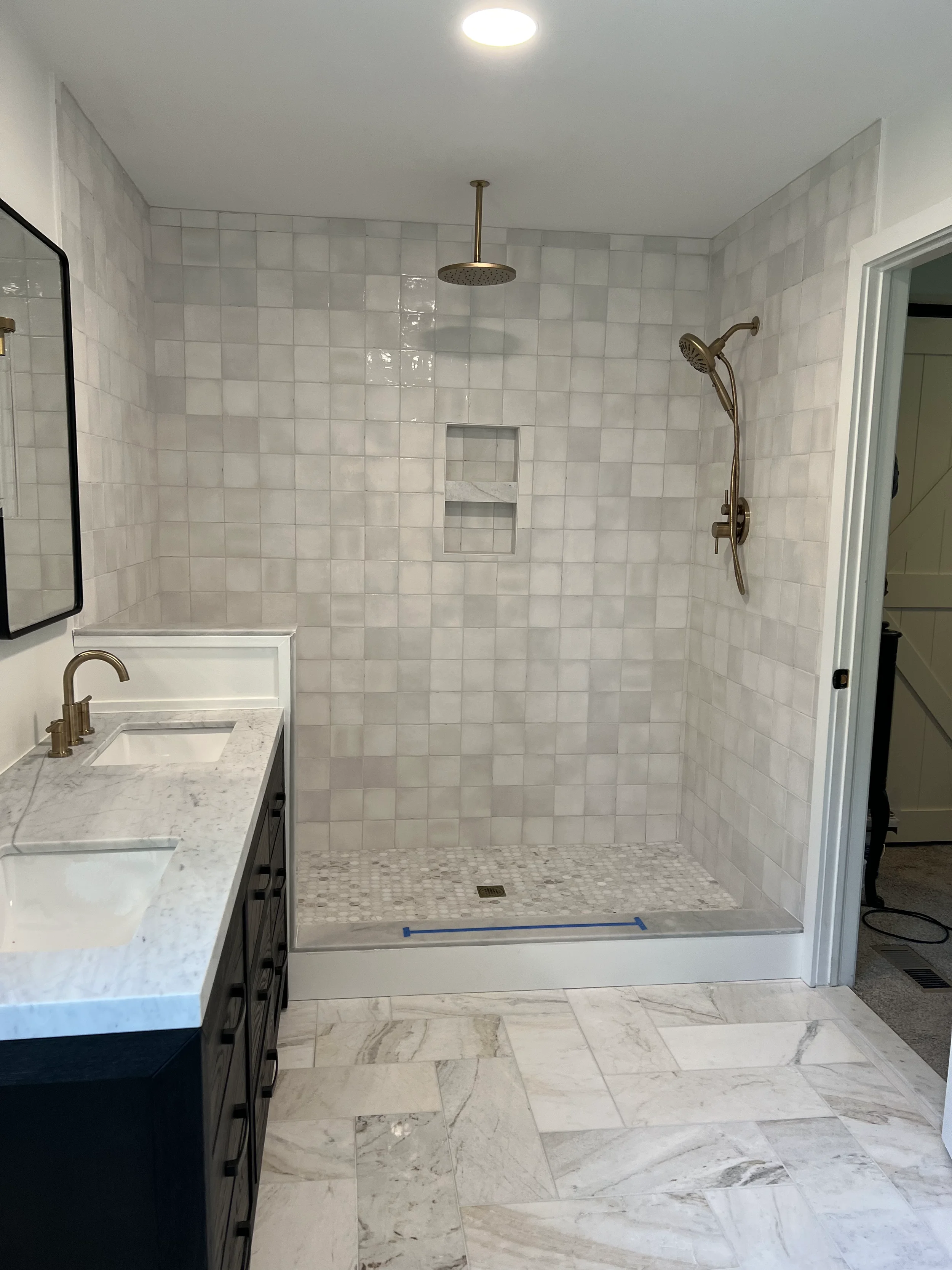 A modern bathroom with a large, open walk-in shower area that has gray square tiles on the walls and marble tiles on the bathroom floor. The shower has a ceiling-mounted rainfall showerhead and a handheld showerhead on the right wall. There is a small built-in recessed shelf in the shower wall. To the left, there is a dark-colored vanity with a white marble countertop, a rectangular undermount sink, and a gold faucet. A mirror with a black frame is partially visible on the wall above the vanity. The bathroom has a bright ceiling light and white trim around the door.