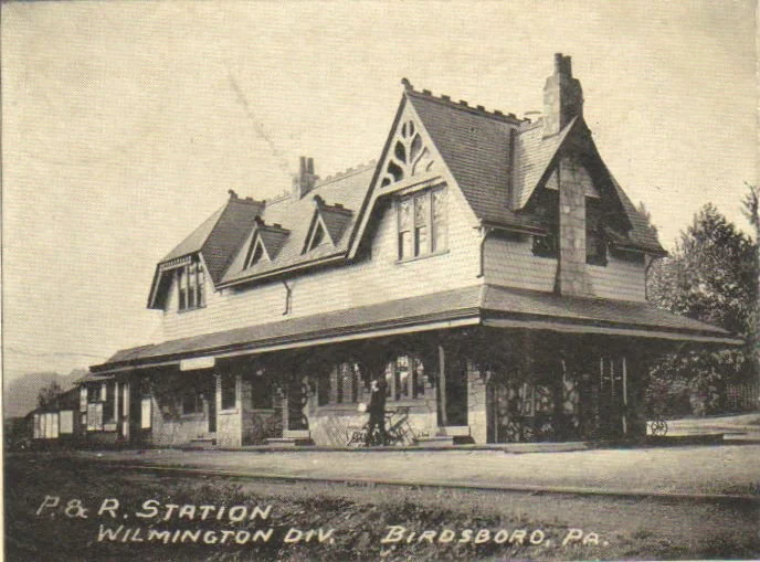 A vintage black-and-white photo of P. & R. Station in Wilmington Division, Bardsboro, Pennsylvania, showing a large, historic train station building with a steeply pitched roof and various gables, supported by brick columns and surrounded by railroad tracks.
