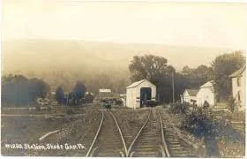 Black and white photo of old train tracks leading toward buildings in a rural area, with trees and open land in the background.