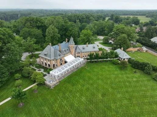 A large historic mansion with a glass-enclosed greenhouse attached, surrounded by lush greenery and tall trees, with a vast lawn in the foreground.