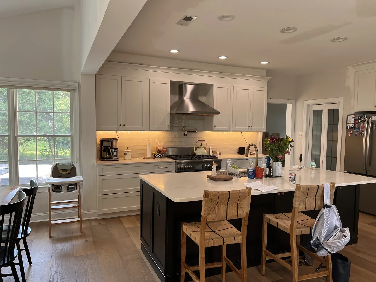 Modern kitchen with white cabinetry, a large kitchen island with a white countertop and black base, stools, stainless steel appliances, including a range with a hood, and a window overlooking greenery.