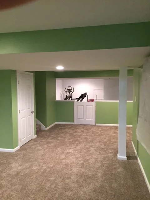 Empty green-walled basement room with beige carpet, white doors, and exercise equipment on a shelf along the back wall.
