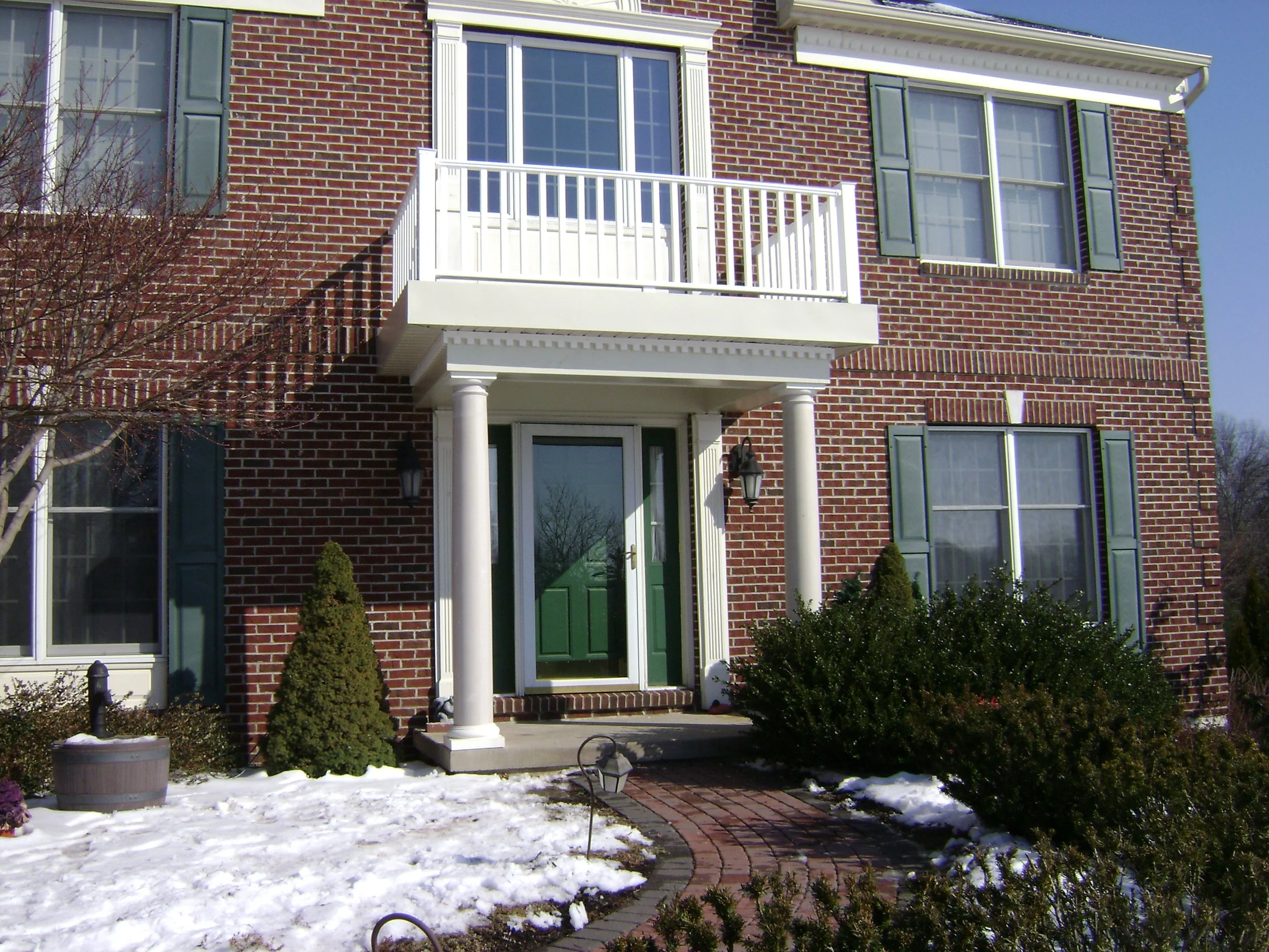 Front view of a brick house with large windows, green shutters, a small balcony, and a porch supported by white columns. The yard has small bushes, a brick pathway, a snow-covered lawn, and outdoor lamps.