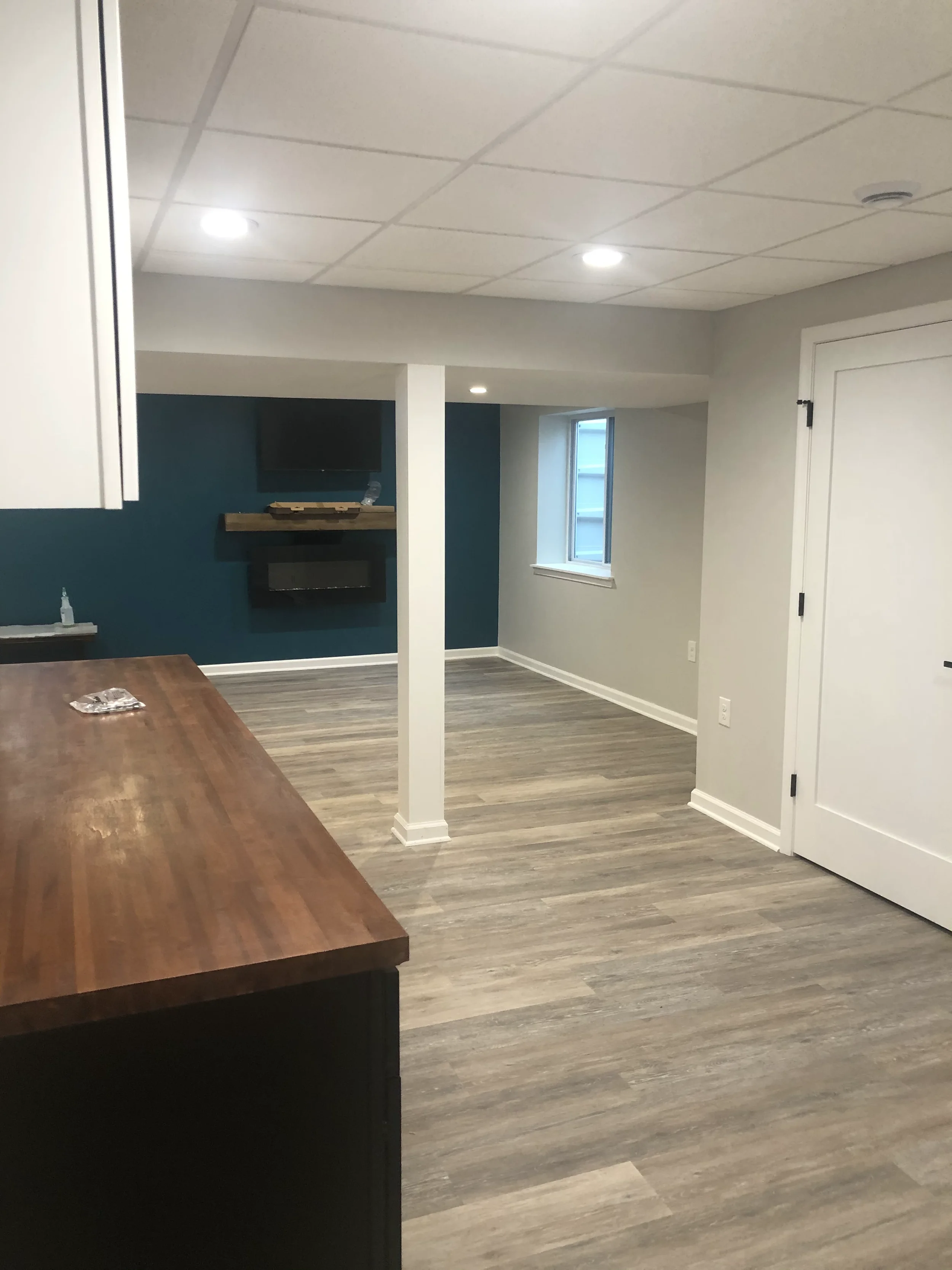 Modern living room with white walls, a blue accent wall, a window, a mounted flat-screen TV, a wooden shelf under the TV, a fireplace, and a ceiling with recessed lighting. There is a wooden dining table in the foreground.