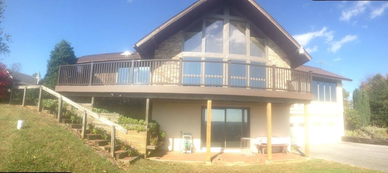 A two-story house with a stone facade and a large upper balcony with glass doors and railing, set against a bright blue sky with some clouds. The house has a sloped roof, large windows, and a backyard with a staircase leading to the yard. There are some plants, a bench, and outdoor items under the house's lower level.