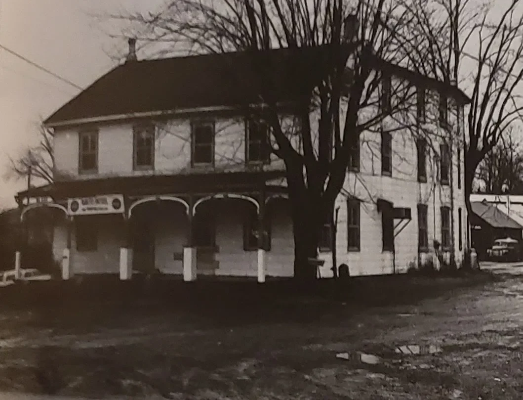 A black and white photo of a two-story house with a porch, several windows, and a large tree in front, with a dirt driveway and a car parked to the right.