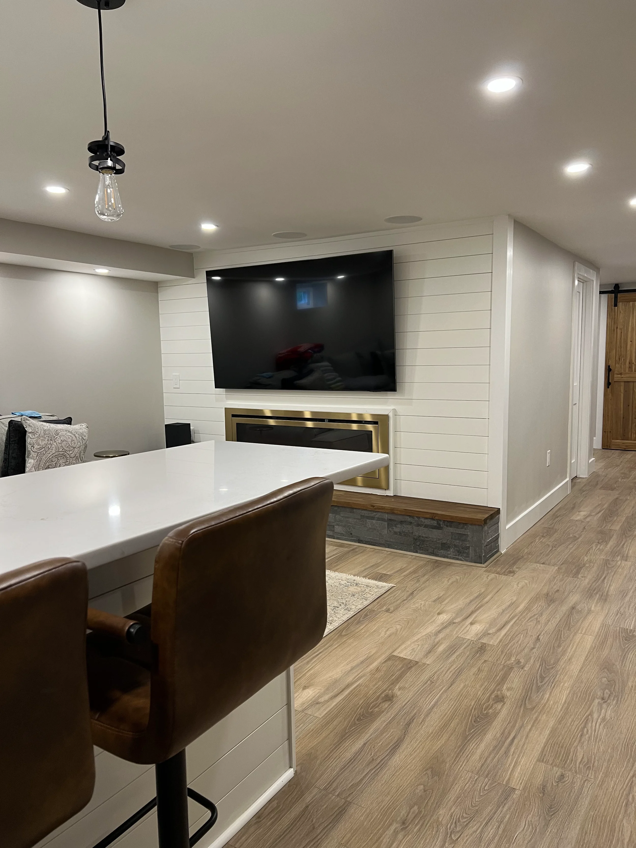 A modern living room with a flat-screen television mounted on a white shiplap wall above a fireplace with a gold trim. There is a white kitchen island with brown barstools in the foreground, and the room has wood flooring and recessed ceiling lights.
