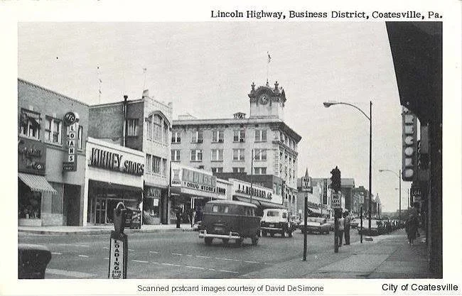 Black and white photo of Lincoln Highway in Coatesville, Pennsylvania, showing storefronts, pedestrians, and cars on the street, with a clock tower building in the background.