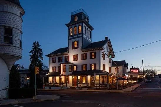 Historic white building with a tower, illuminated at dusk, on a street corner with outdoor seating and neighboring structures.
