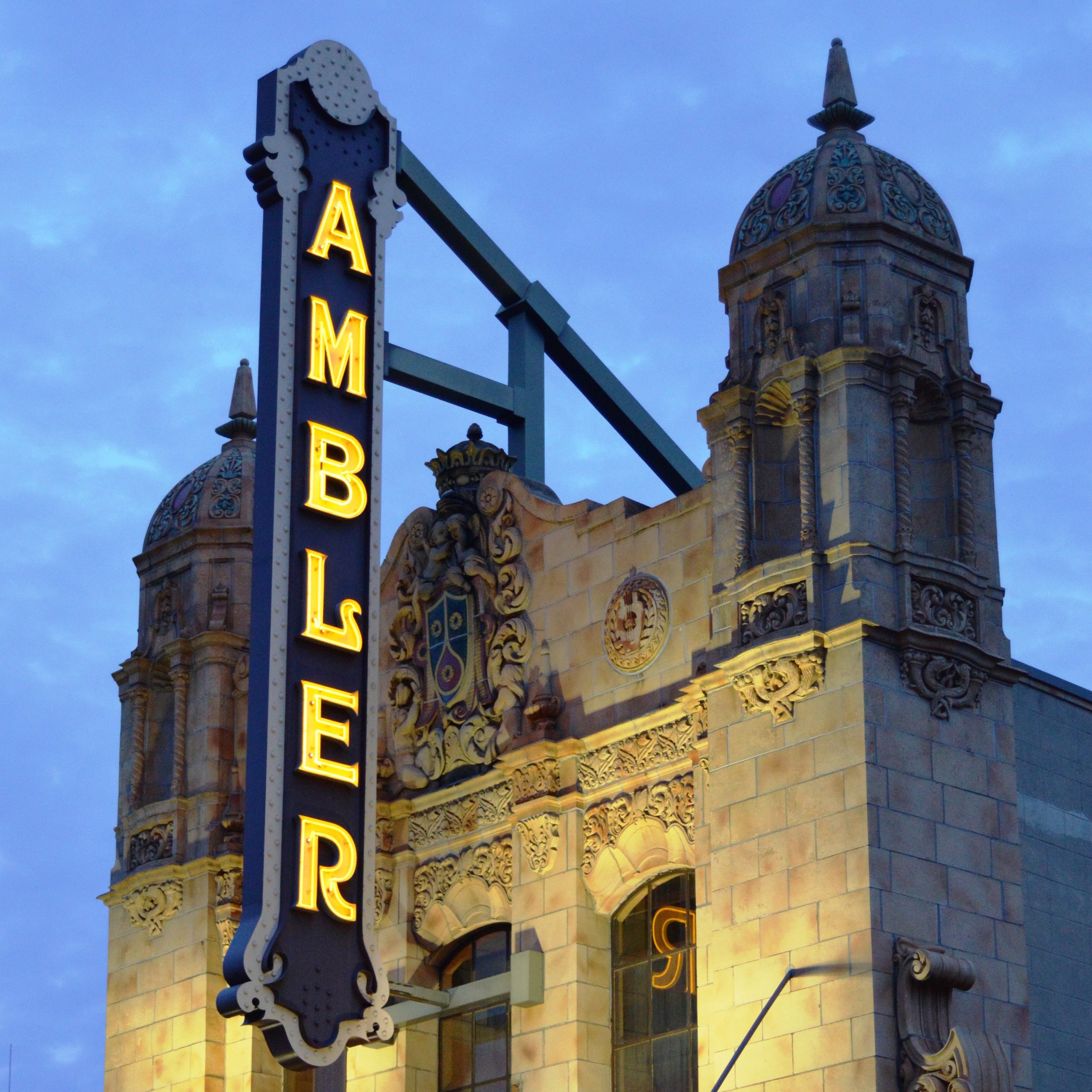 Front corner of a historic building with ornate architecture, featuring a sunset sky in the background. There is a vertical sign with yellow illuminated letters spelling 'AMBER'. The building has a decorative dome with intricate design and a small spire on top.
