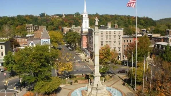 An aerial view of a town square with a monument at the center, surrounded by trees, buildings, and flagpoles with American flags.