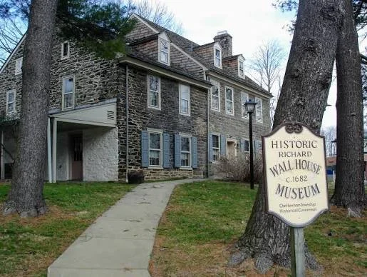 Historic Richard Wall House Museum, a stone building with multiple windows and blue shutters, situated behind a sidewalk and trees.