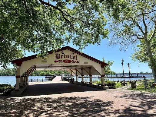 Picture of the Bristol Pavilion by a lake, with trees, benches, and a clear blue sky.