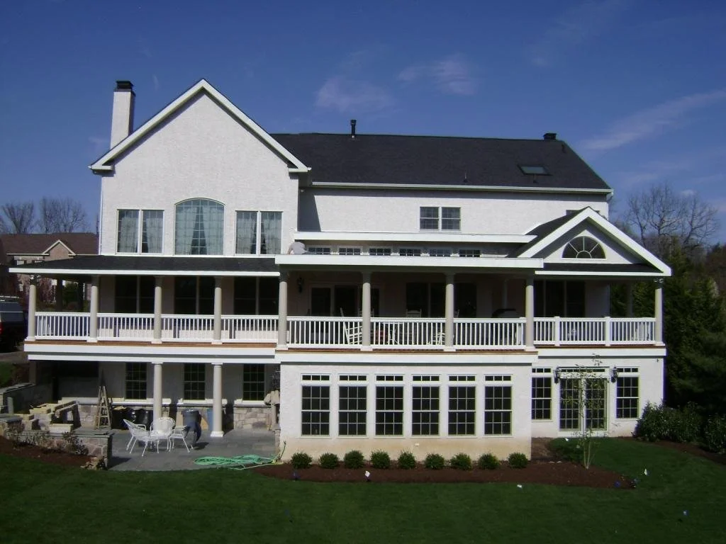 Large white multi-story house with balconies, large windows, and a sloped roof, set against a blue sky with some trees in the background.