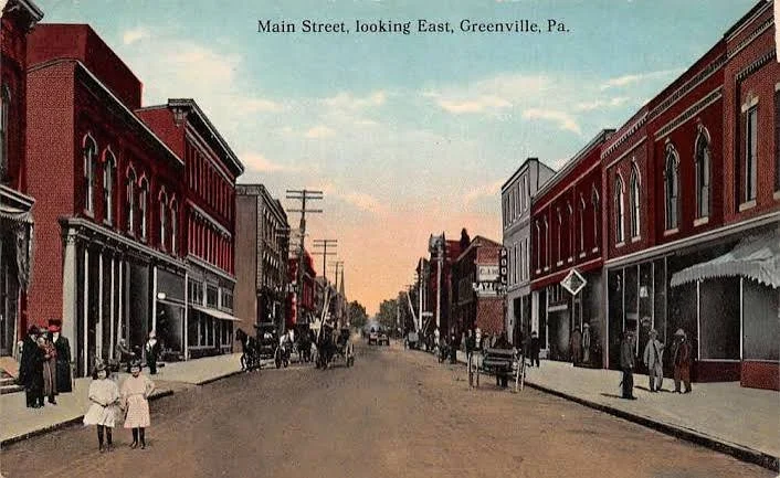 Main Street in Greenville, Pennsylvania, looking east, with brick buildings and pedestrians, some in early 20th-century attire, and horse-drawn carriages on the street at sunset.