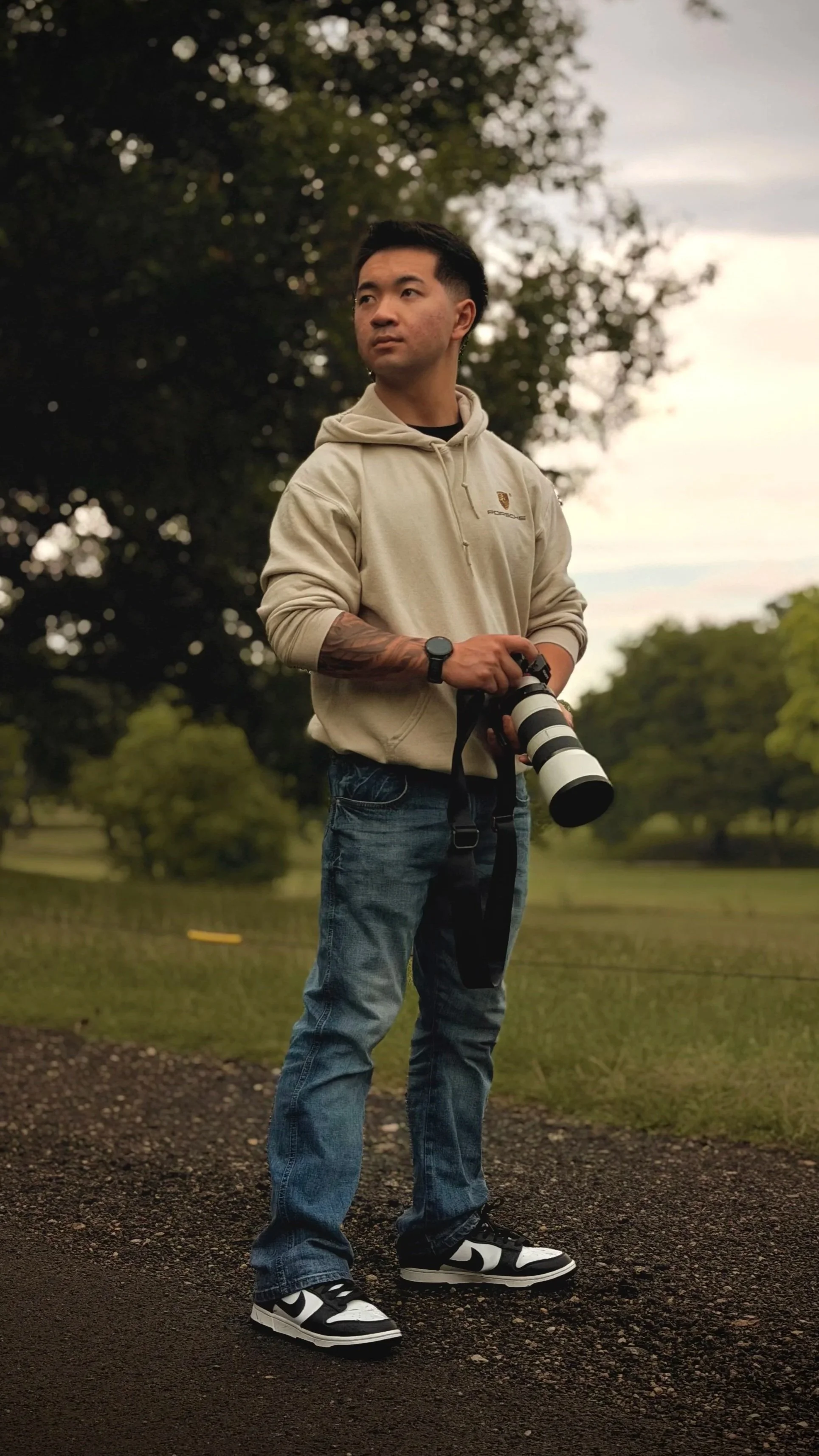 A young man with dark hair and tattoos on his arm standing outdoors on a trail, holding a professional camera with a large telephoto lens, wearing a beige hoodie, jeans, and black-and-white sneakers, with trees and a cloudy sky in the background.