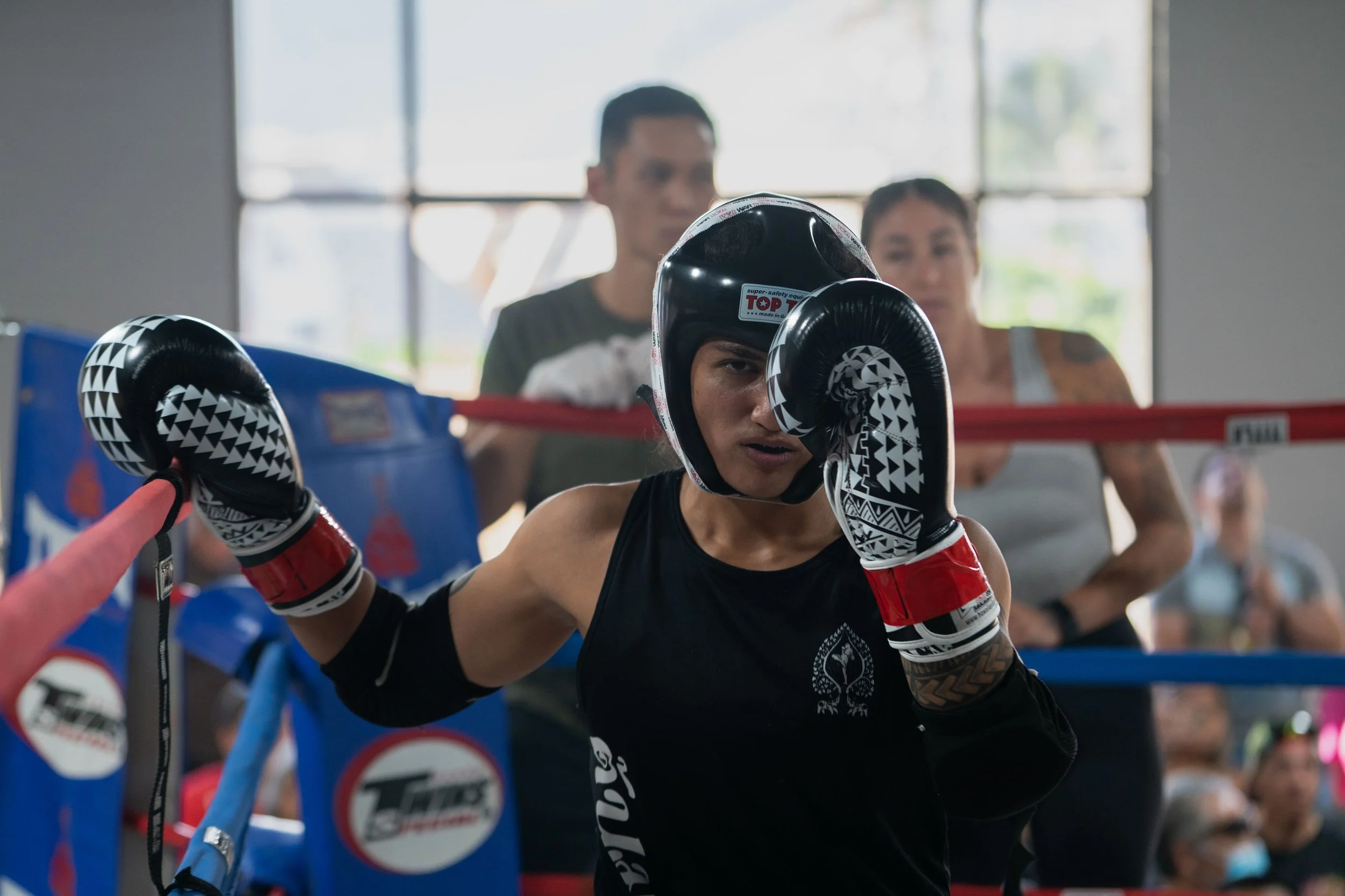 A female boxer in black gloves and headgear sparring in a boxing gym, with a male and female spectator watching behind her.
