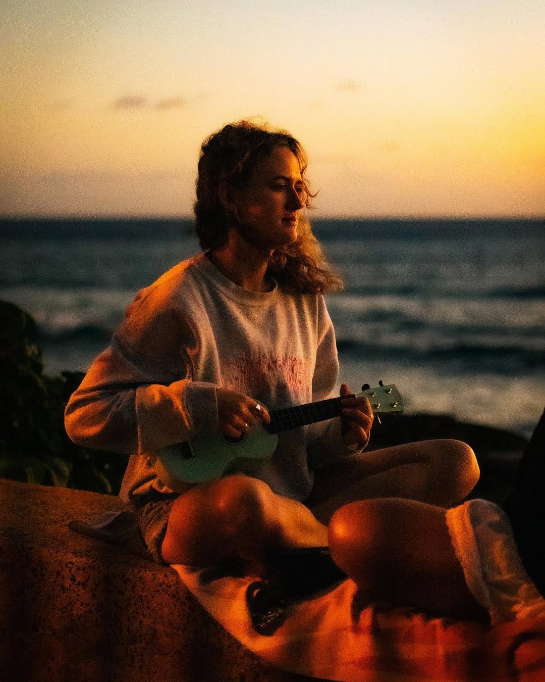 A young woman sitting on a blanket by the beach during sunset, playing a small ukulele.