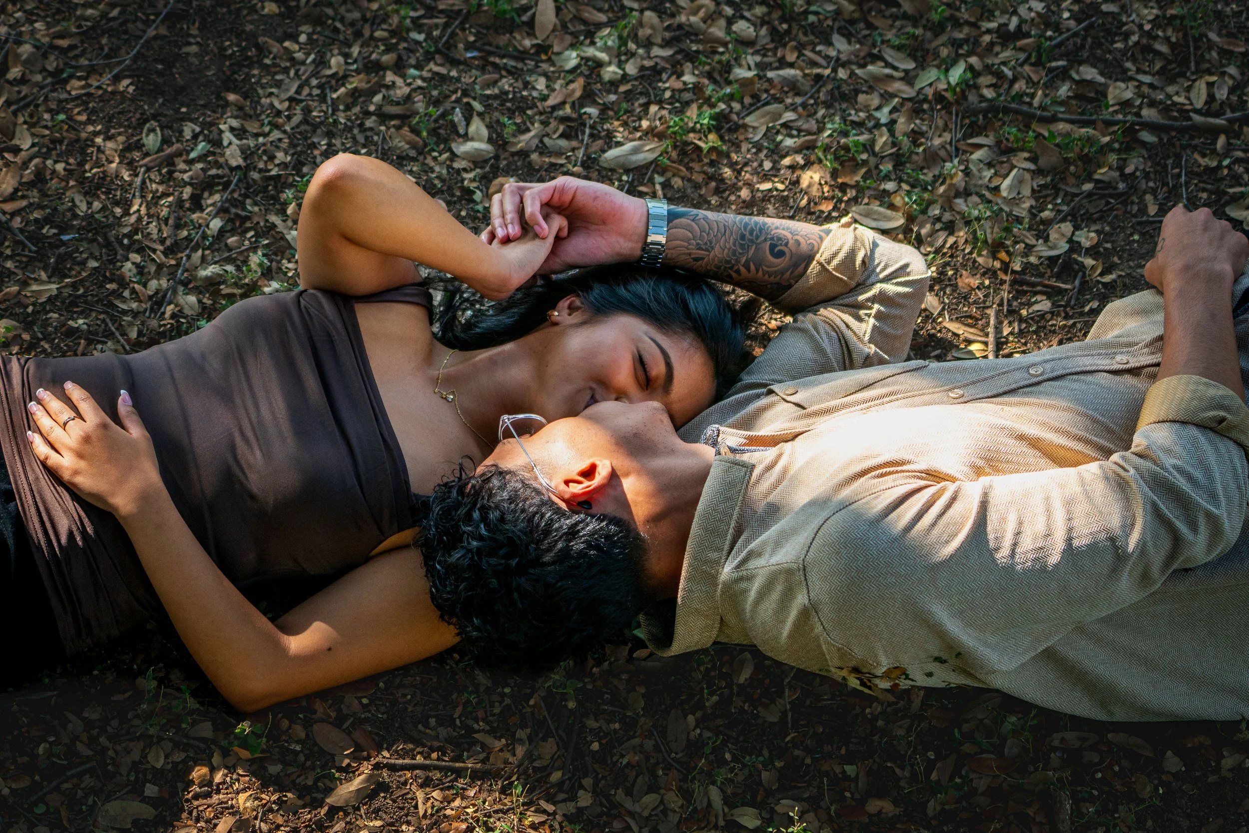 Two women and a man lying on the ground outdoors, eyes closed, holding hands and sharing a tender moment.