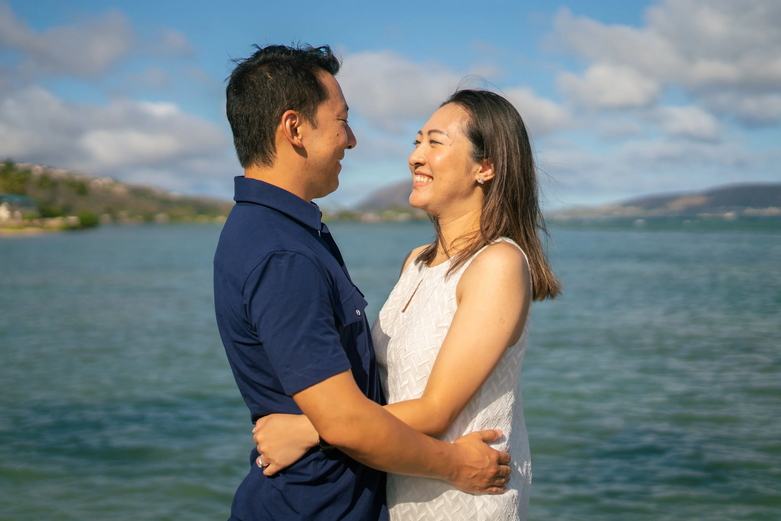 A couple standing close together on a body of water, smiling at each other with a scenic landscape and blue sky in the background.