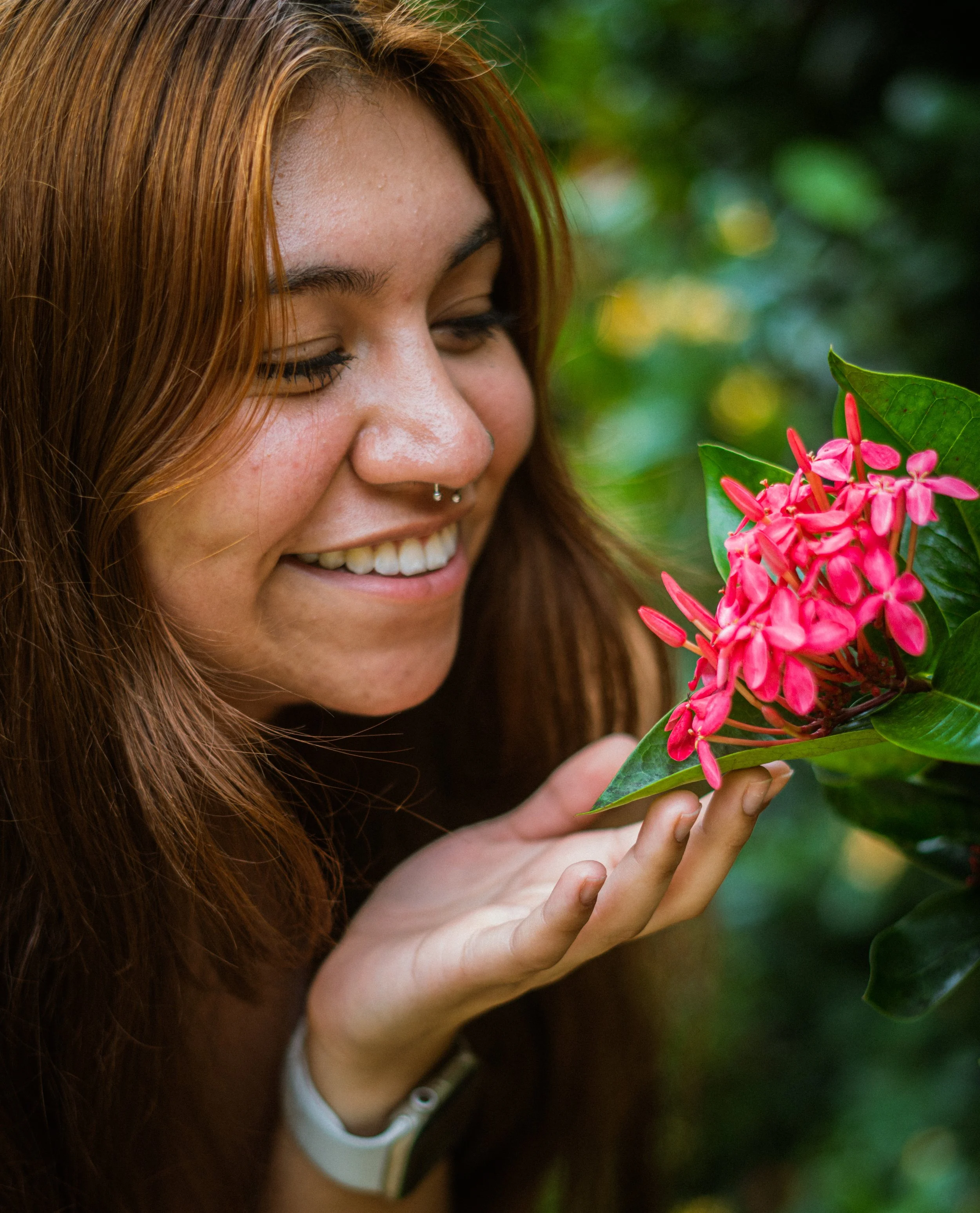A woman with long red hair smiling while holding pink flowers with green leaves.