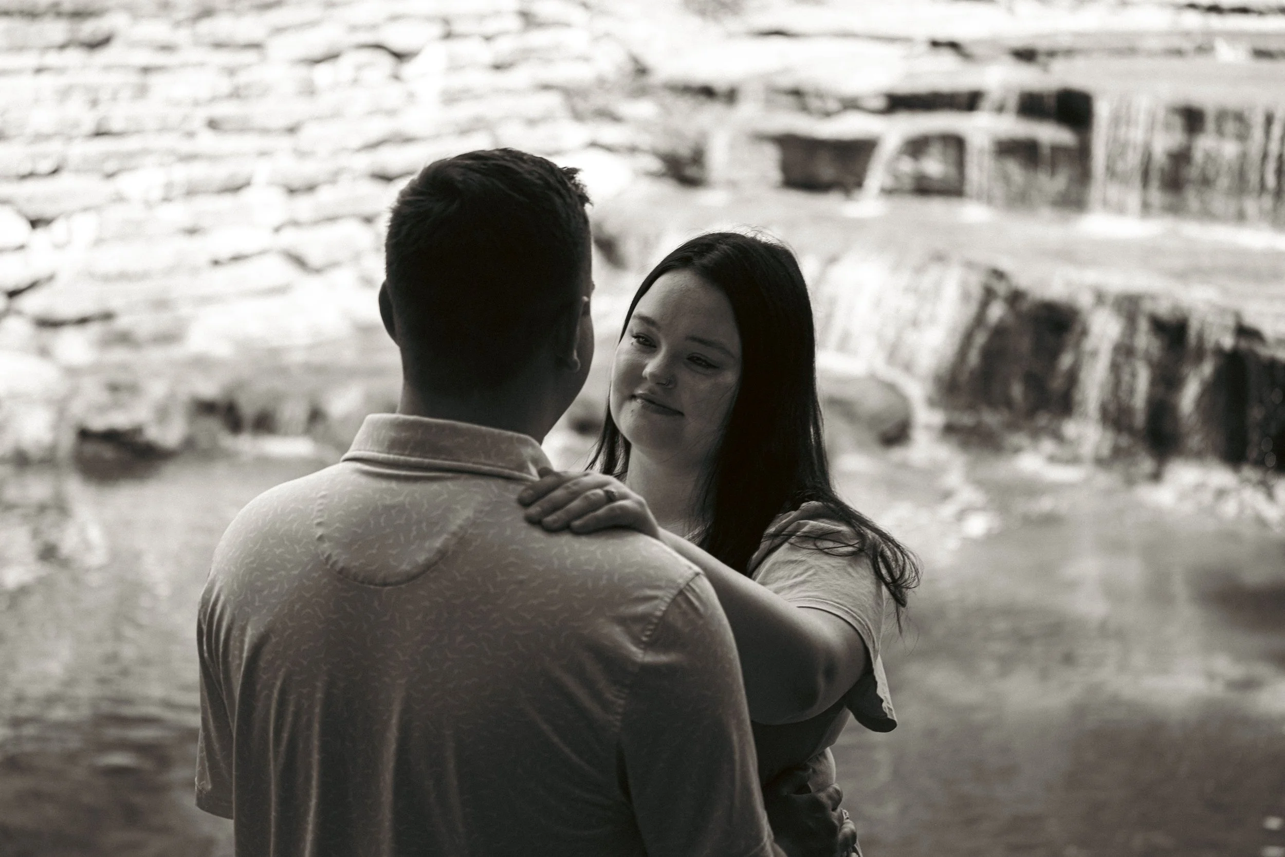 A couple, a man and a woman, dancing closely outdoors near a body of water with a rocky backdrop, engaged in an intimate moment.