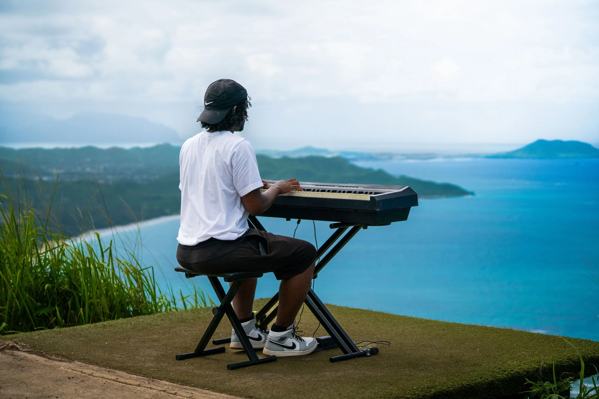 A person wearing a black cap, white t-shirt, and Nike sneakers sitting on a foldable stool and playing a keyboard outdoors near the ocean on a cloudy day.