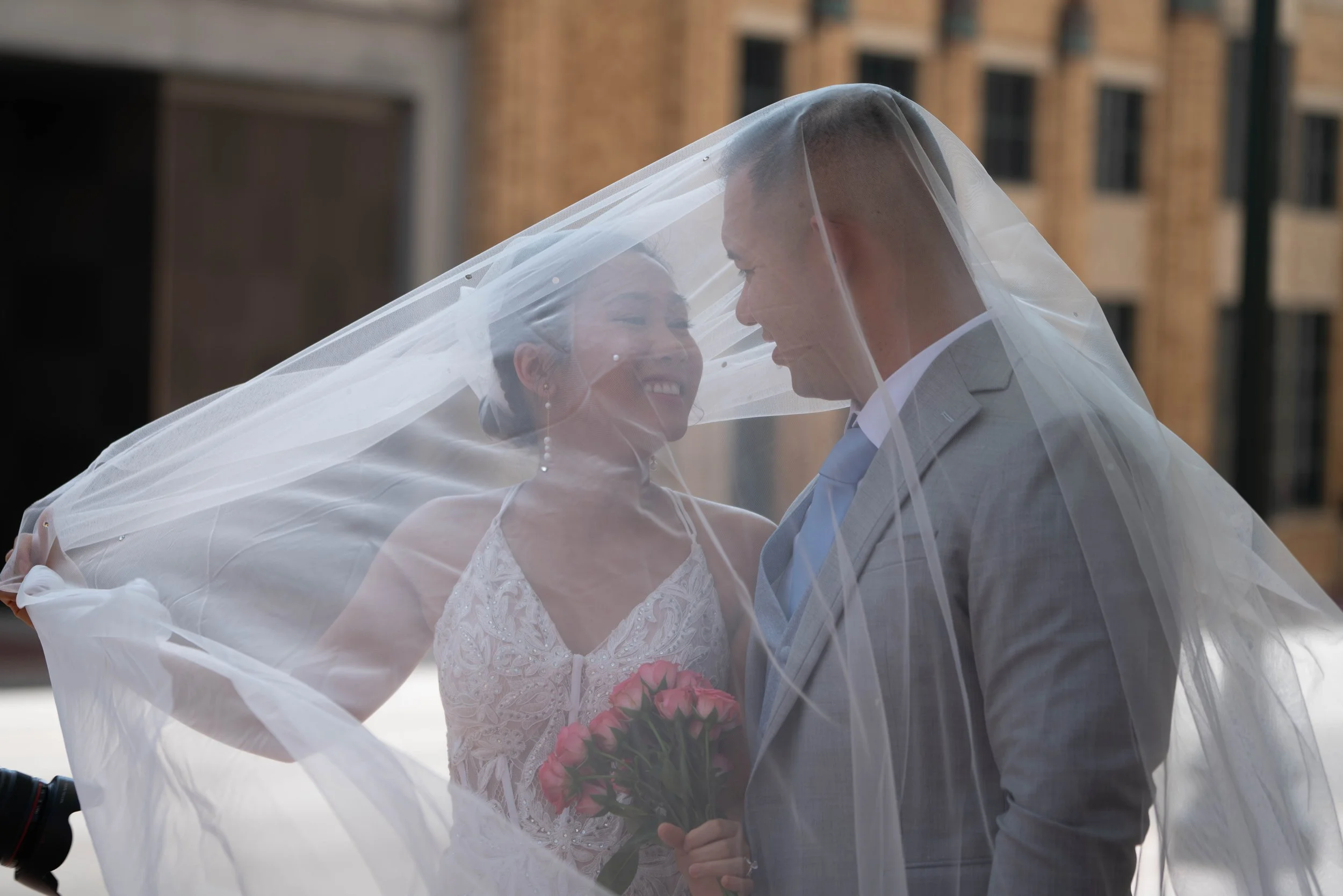 A bride and groom smiling and looking at each other under a sheer white veil, with the bride holding a pink bouquet, outside in front of a brick building.
