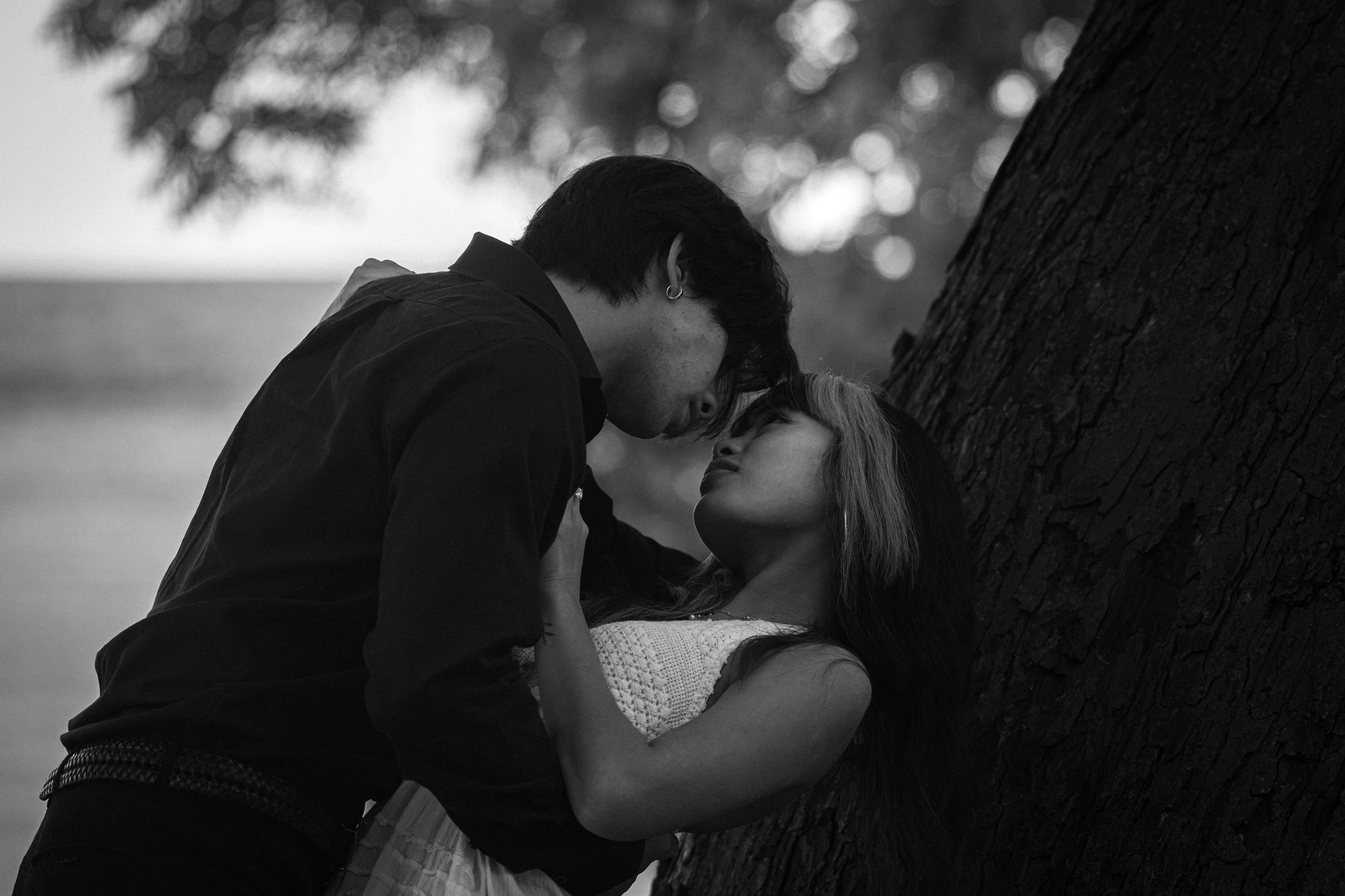 A black and white photo of a couple close to each other outdoors, with their faces near and eyes closed, sharing an intimate moment next to a large tree.
