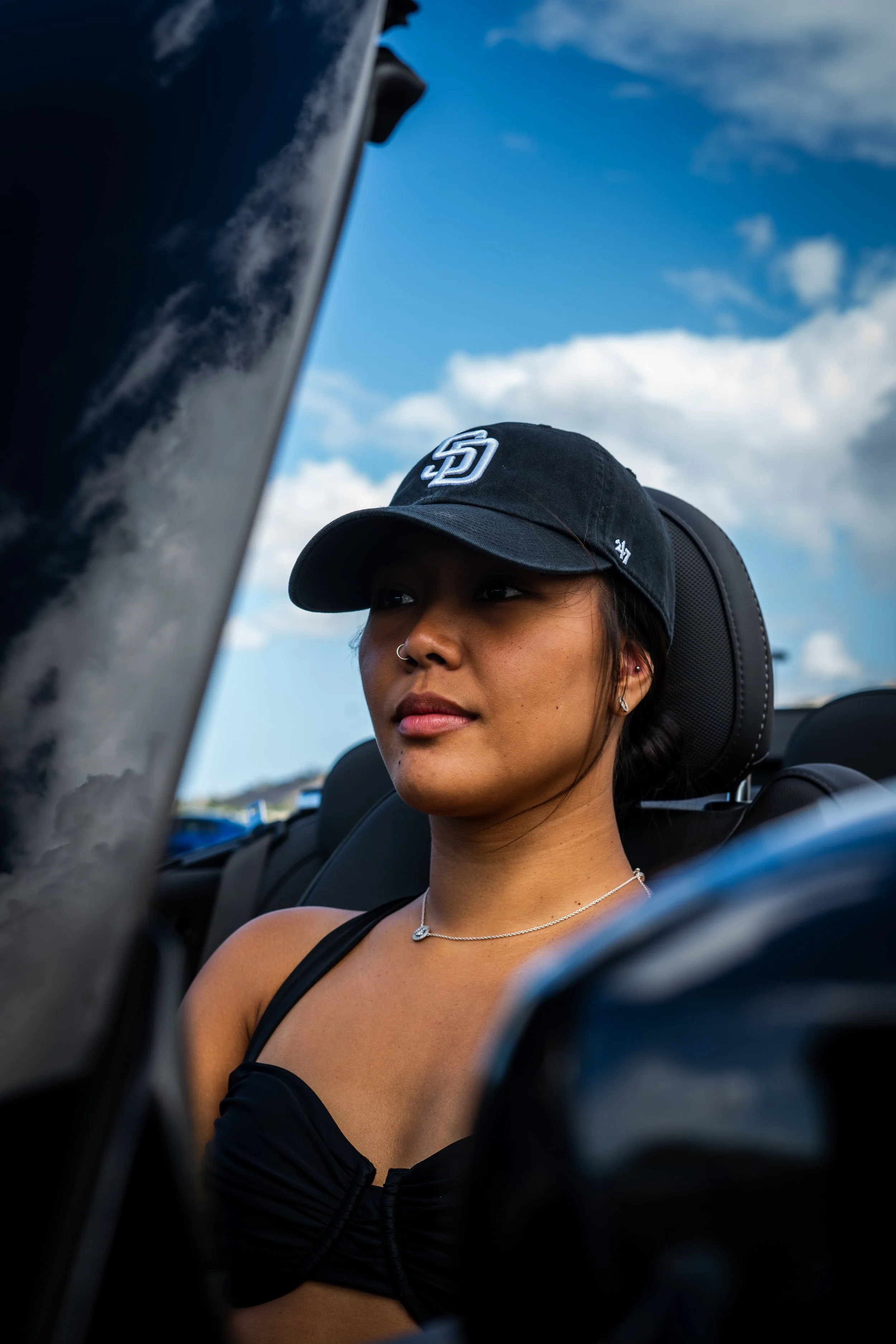 A woman with a dark baseball cap, necklace, earrings, and a nose ring sitting inside a vehicle with a blue sky and white clouds visible outside.