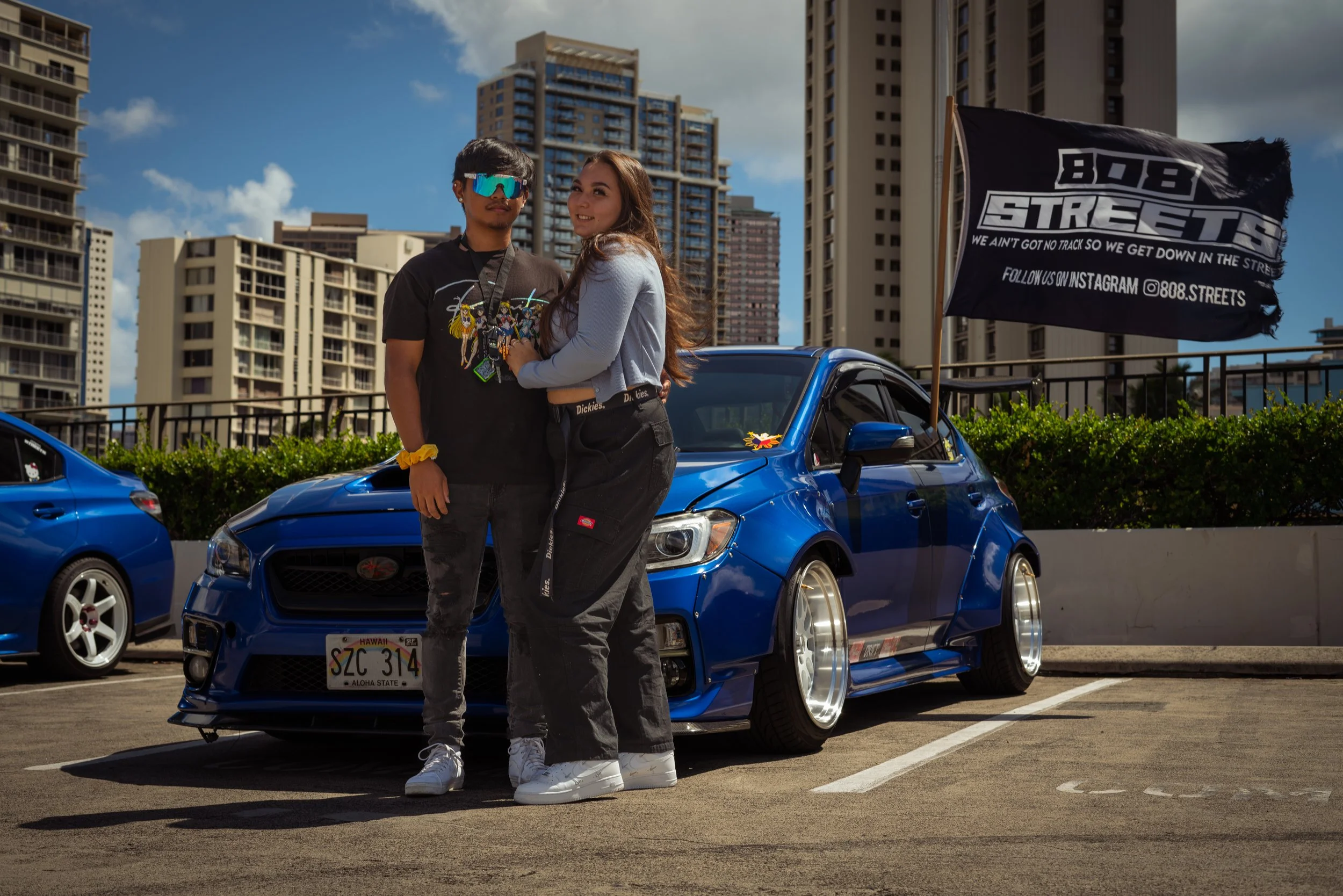 Two young people, a man and a woman, standing close together in a parking lot in front of a blue sports car. They are outdoors with high-rise buildings in the background. The man is wearing sunglasses, a graphic T-shirt, and dark pants. The woman is 