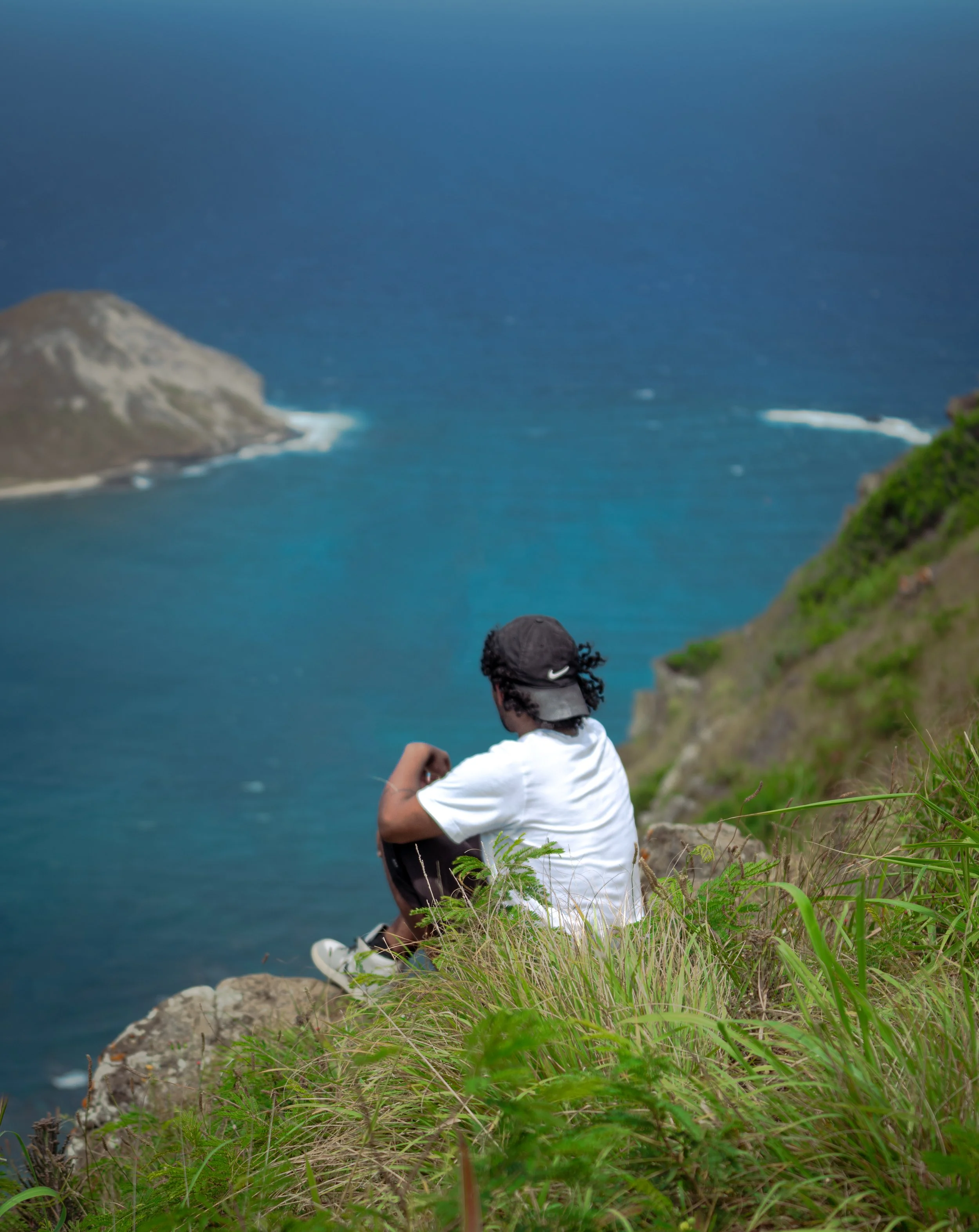 A person sitting on a grassy cliff overlooking the ocean, with a view of the coast and water, wearing a white shirt and a black cap.