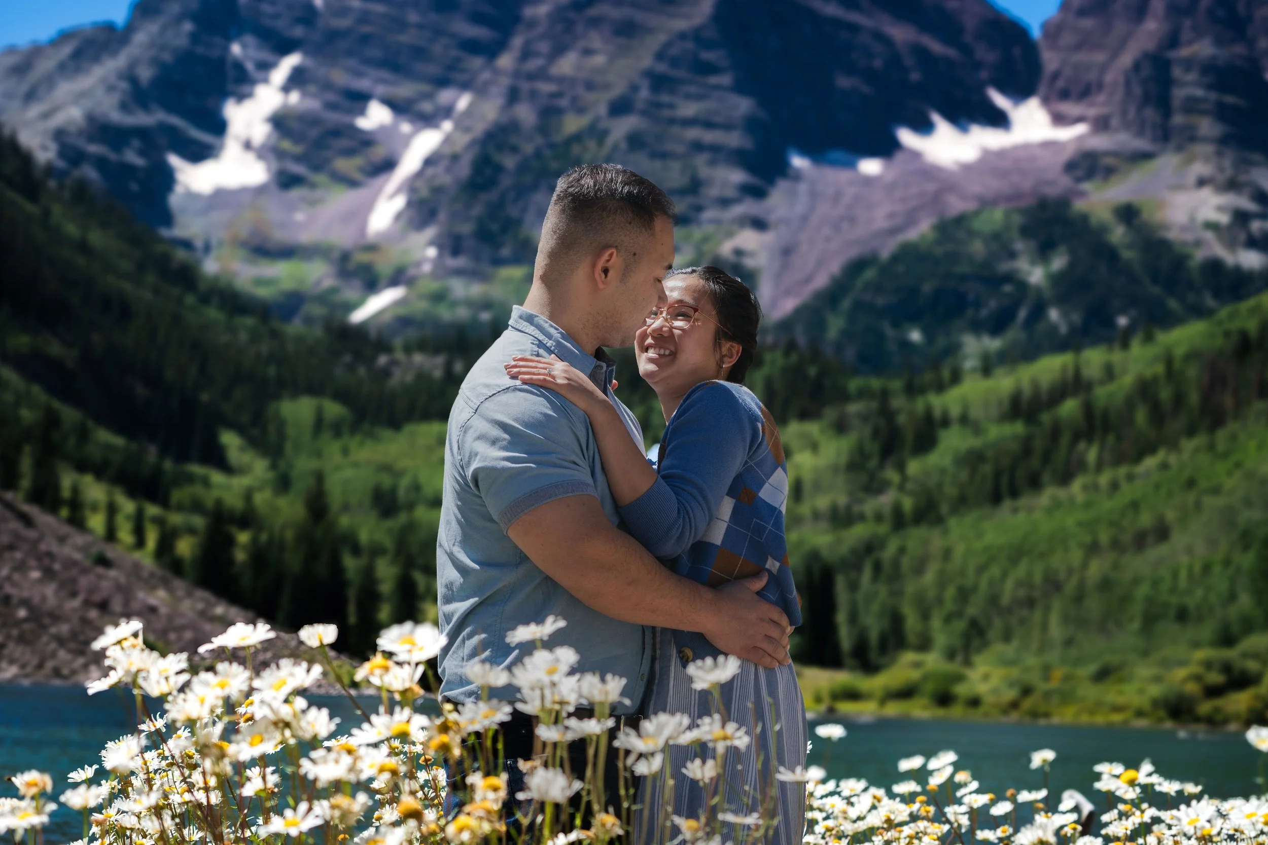 A couple embracing outdoors near a lake with mountains in the background, surrounded by white daisies.