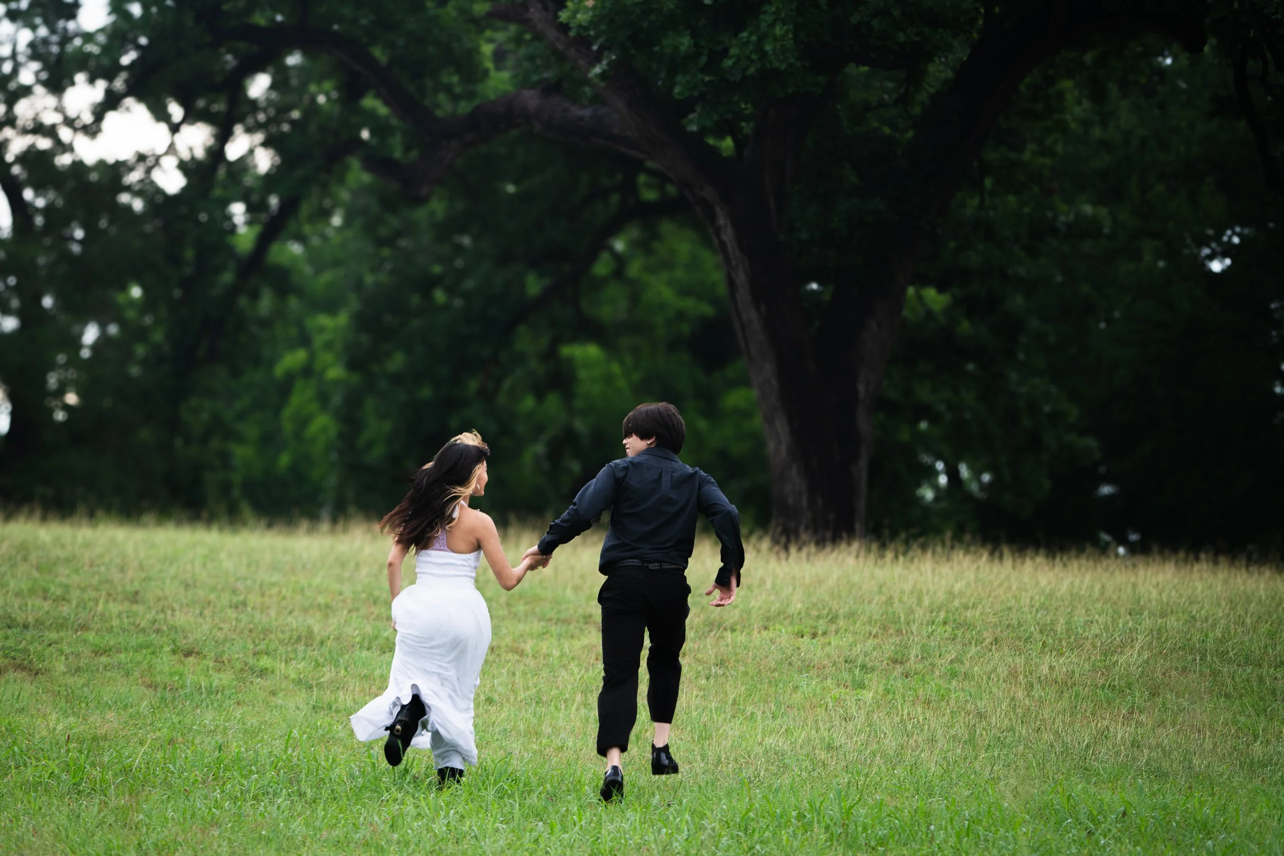 A young girl in a white dress and a young boy in black pants holding hands and running through a grassy field with large trees in the background.