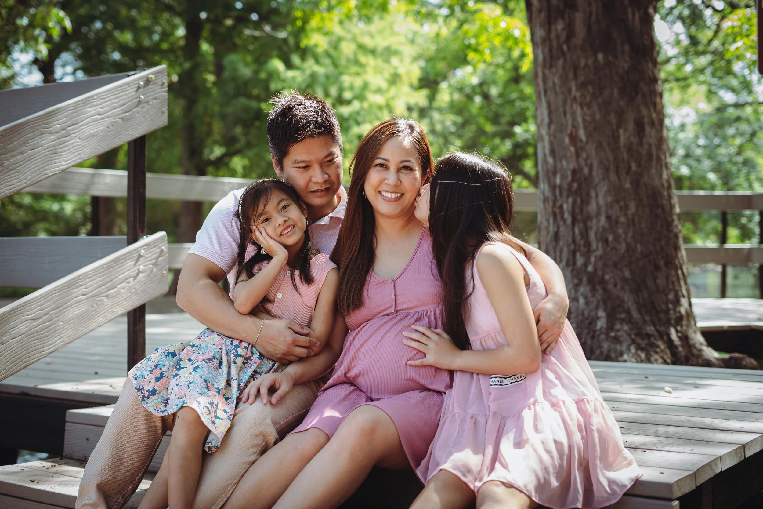 A family of five sitting on a wooden dock outdoors, with trees and sunlight in the background. The family consists of a man, a woman, two girls, and a pregnant woman, all smiling and close together.