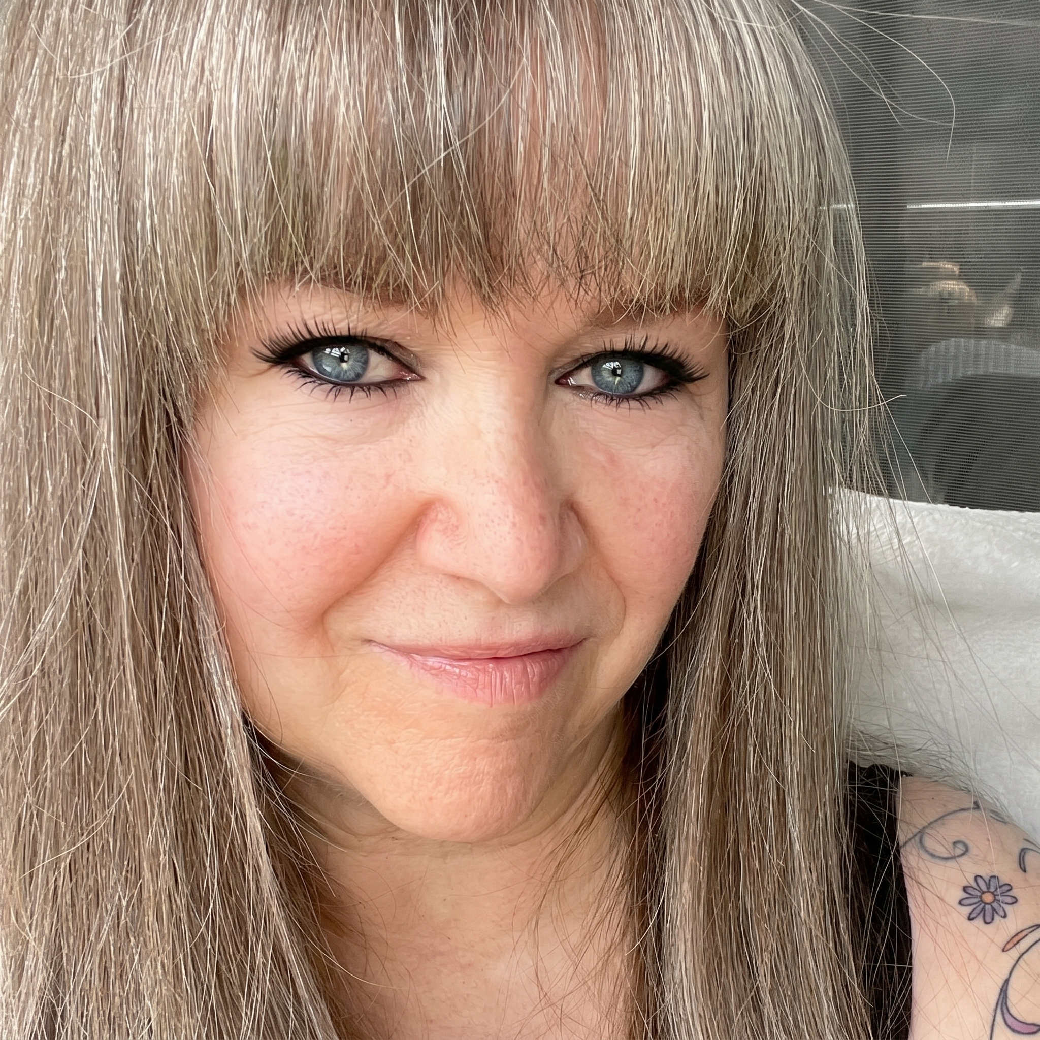 Close-up of a woman with blonde hair, blue eyes, and a floral tattoo on her shoulder. She has a slight smile and is looking at the camera.