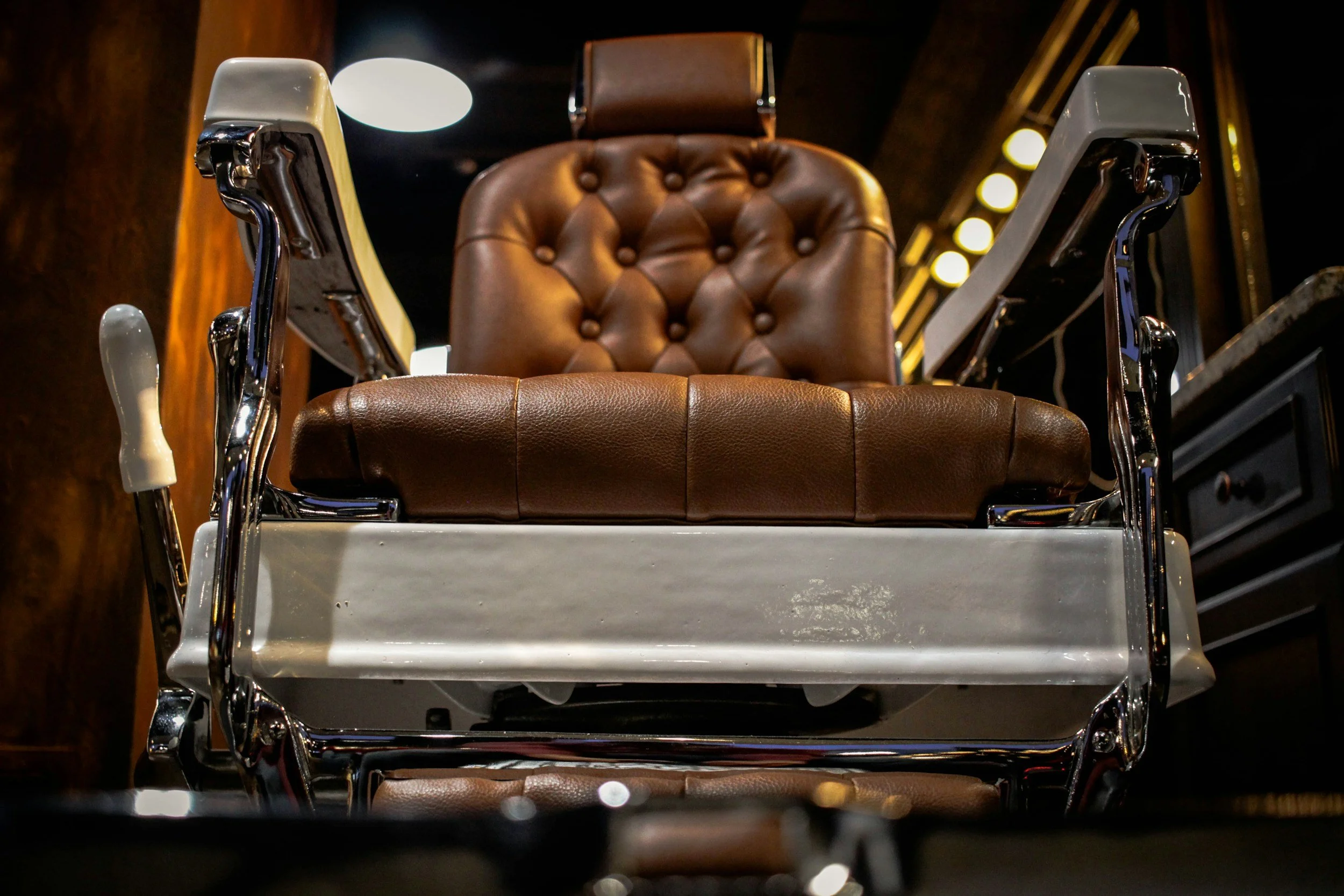 A vintage barber chair with brown cushioned upholstery and shiny metal armrests, positioned in a dimly lit room.