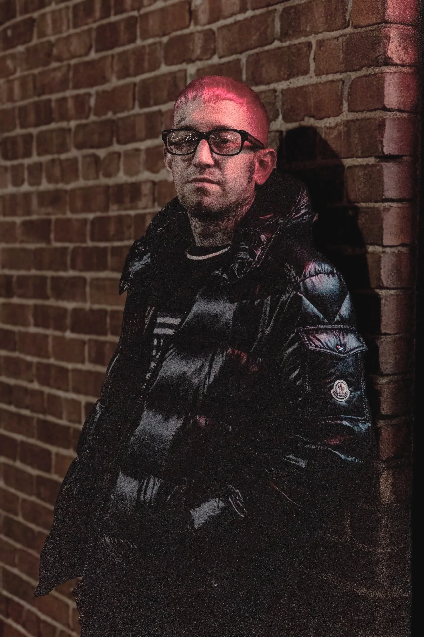 A man with short blonde hair, glasses, and tattoos on his neck, standing against a brick wall at night, wearing a shiny black jacket.