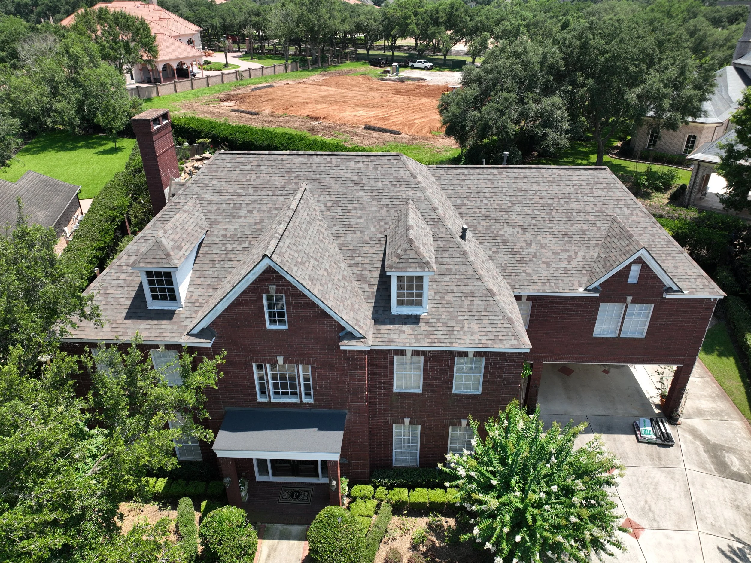 An aerial view of a residential neighborhood showing a large house with a gray shingle roof, surrounded by trees and greenery. Behind the house, there is a cleared dirt lot and a few parked cars.