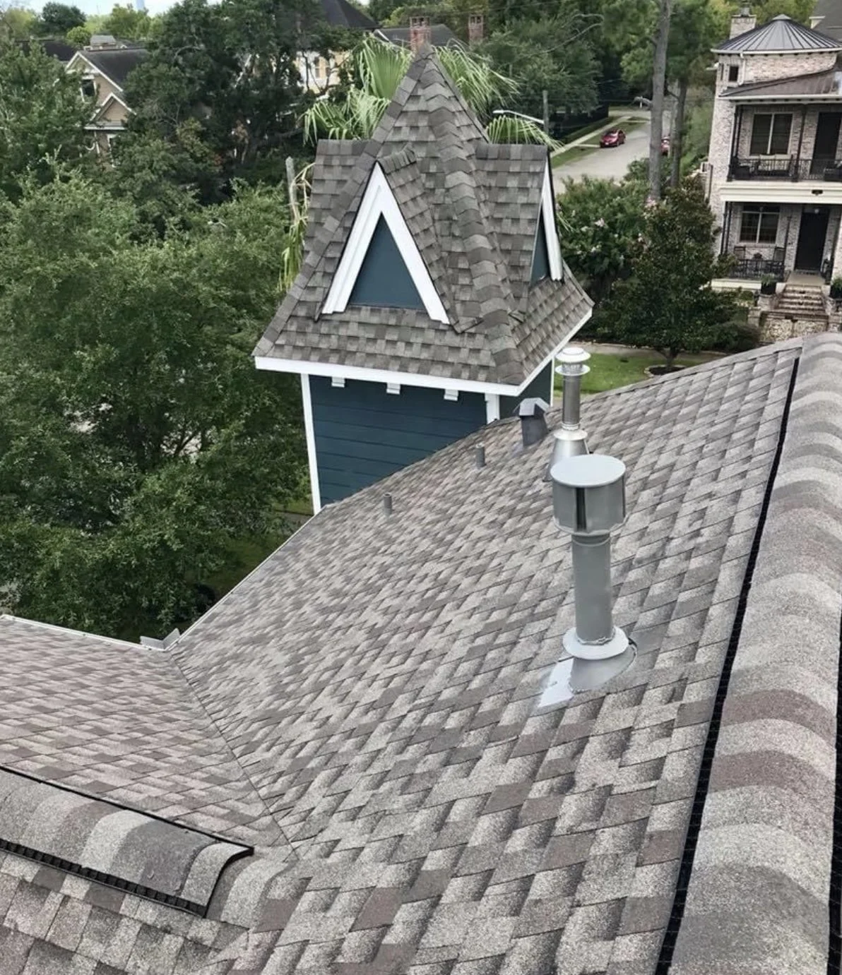 A rooftop with asphalt shingles, visible chimney, and a small weather vane. There is a small blue building with a steep roof and triangular windows, and a background of trees and houses.