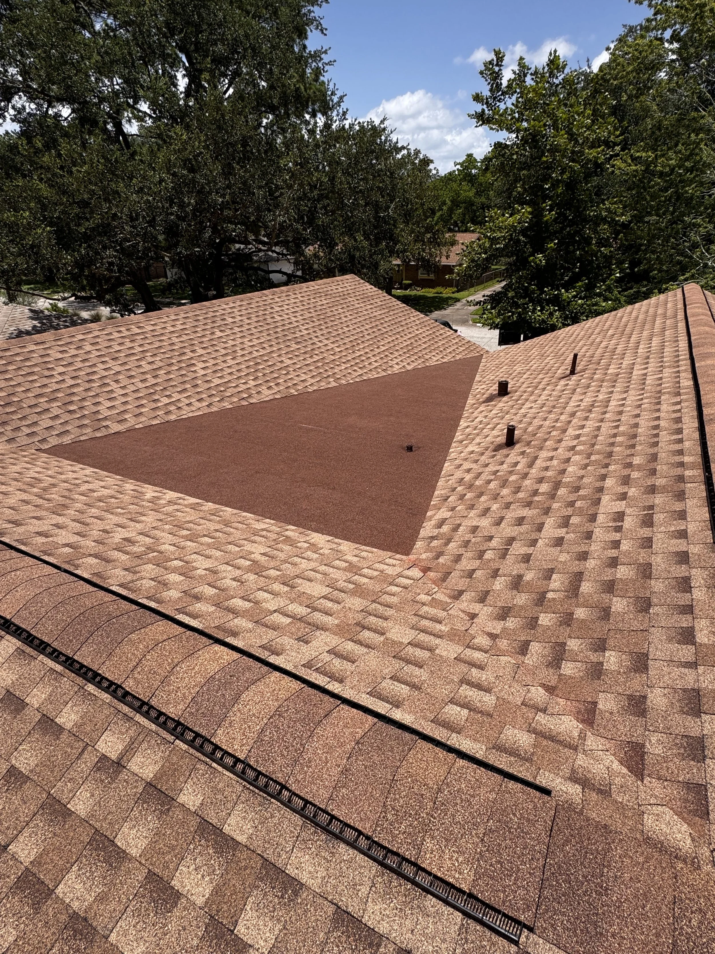 View of house rooftops with asphalt shingles in shades of brown, surrounded by trees with green foliage, under a partly cloudy blue sky.