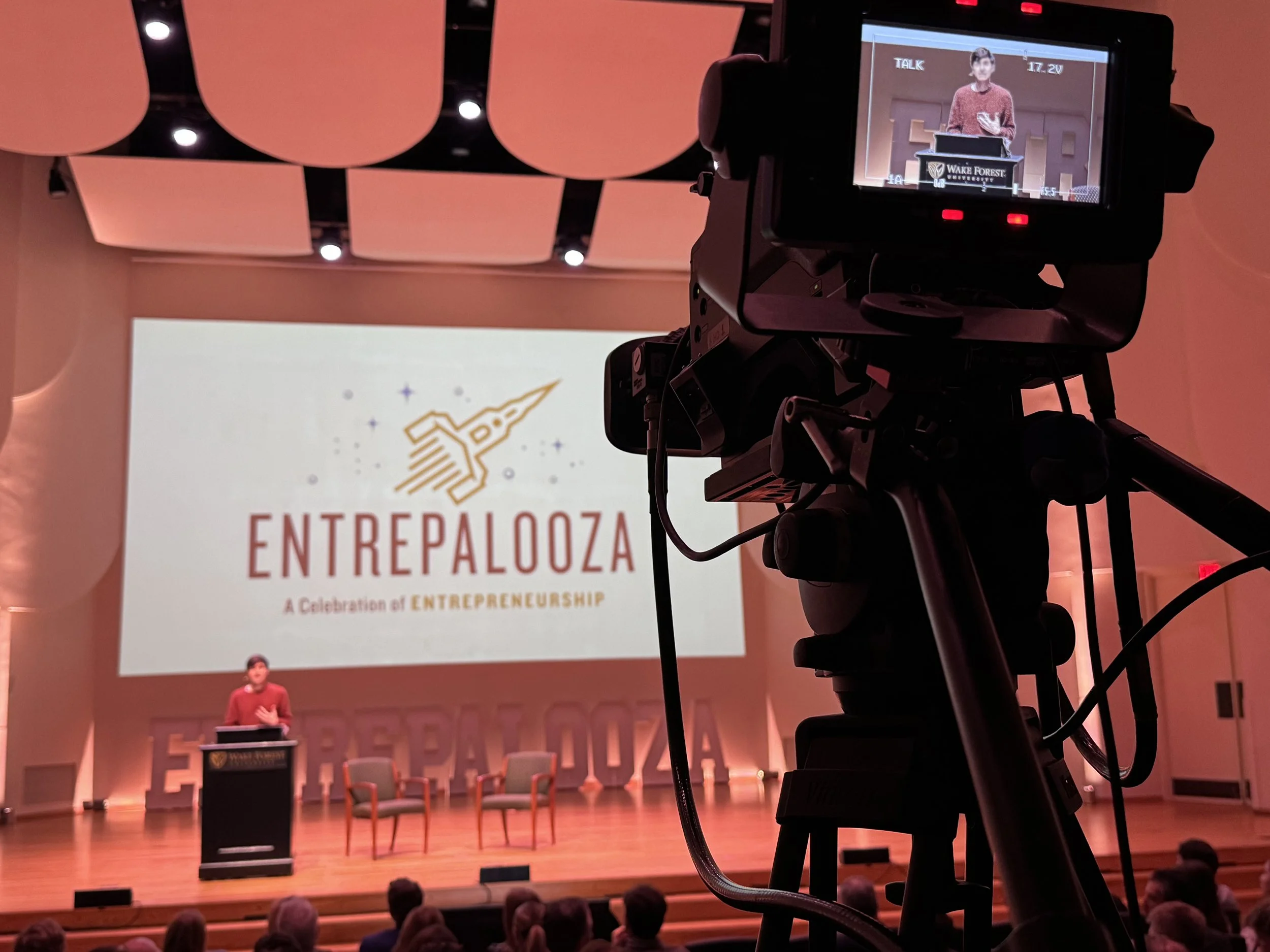 A speaker stands at a podium with the Wake Forest logo, giving a presentation on a stage in front of a large screen displaying the event logo 'Entrelazoza, A celebration of entrepreneurship.' There are chairs on stage, and an audience is seated in fr