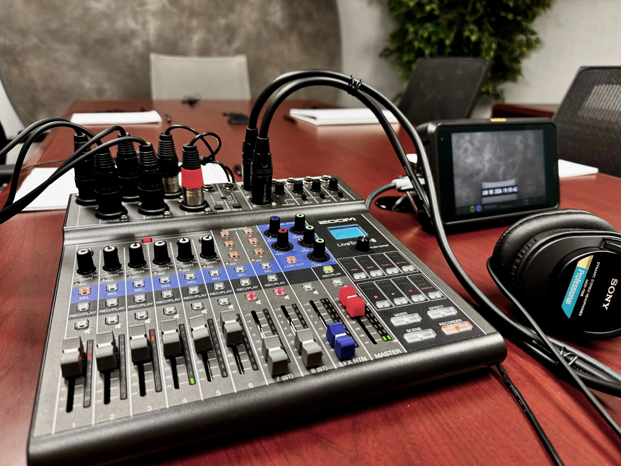 Audio mixing console with various knobs and sliders, connected to a digital audio recorder and headphones, on a wooden table in a conference room.