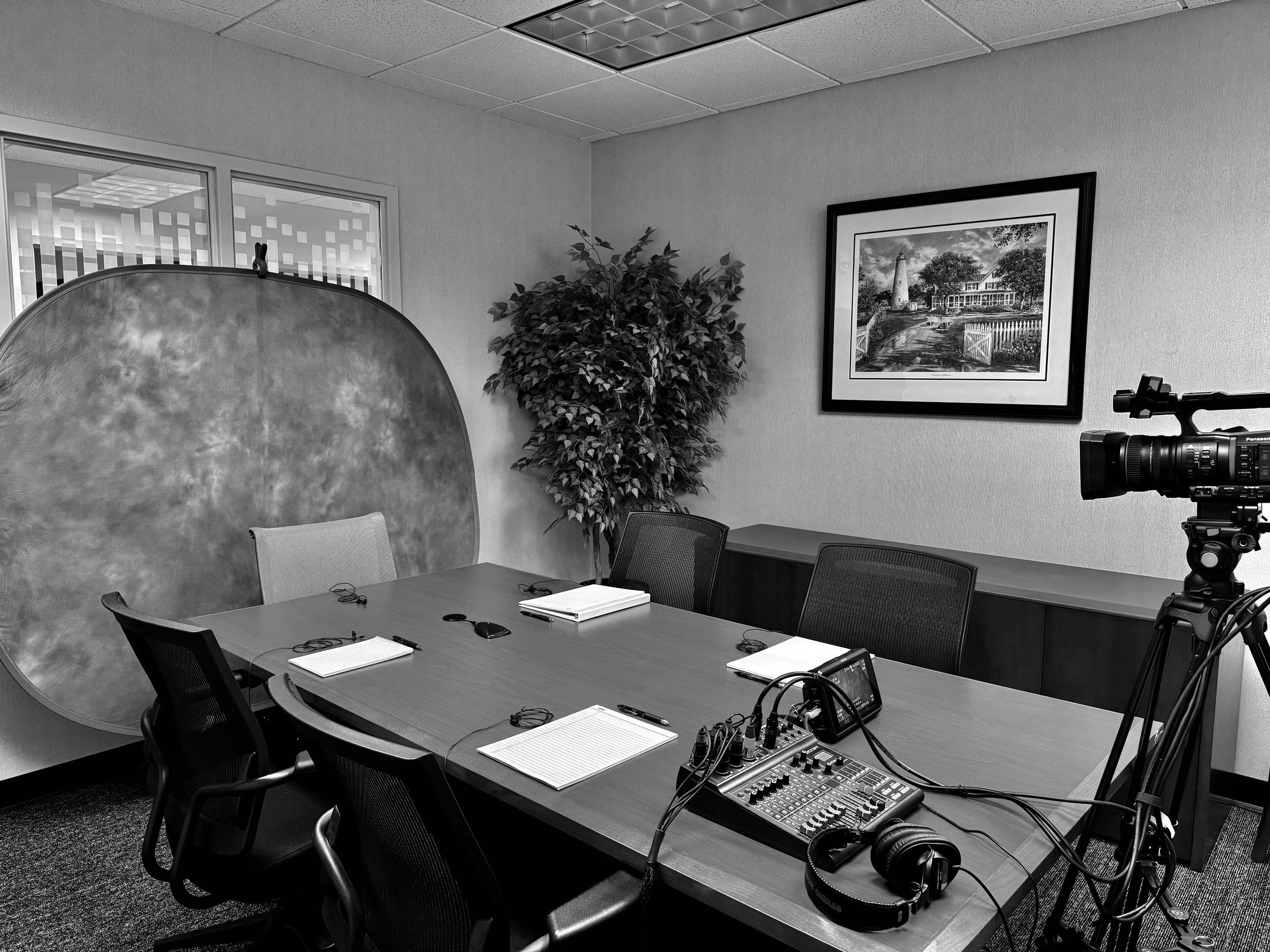 A conference room with a large wooden table surrounded by nine chairs. On the table are notebooks, headphones, a smartphone, and an audio mixer with cables. A large framed black-and-white landscape painting hangs on the wall, which also has a large p