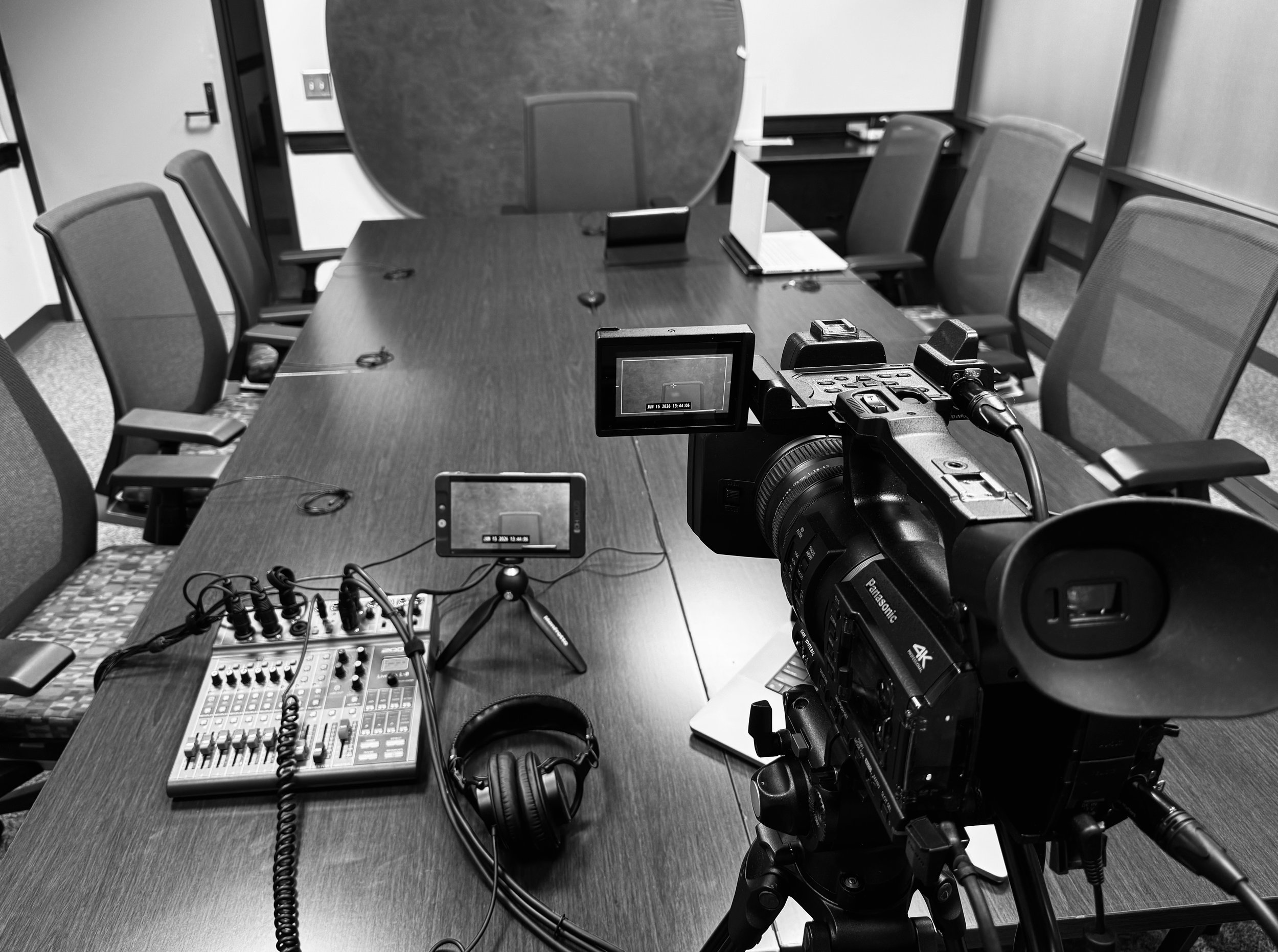A conference room with a long tabletop surrounded by chairs, with video and audio equipment, including a camera, headphones, an online meeting display, and a mixing console, set up for a recording or broadcast.
