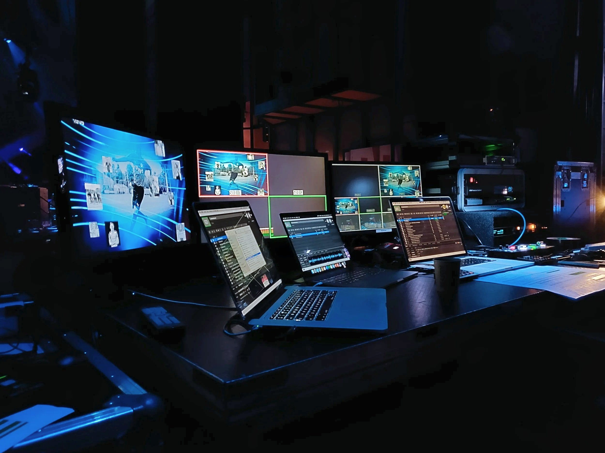 A dark room with multiple screens and laptops displaying live video feeds and controls, indicating a control room, edit suite, or broadcast setup.
