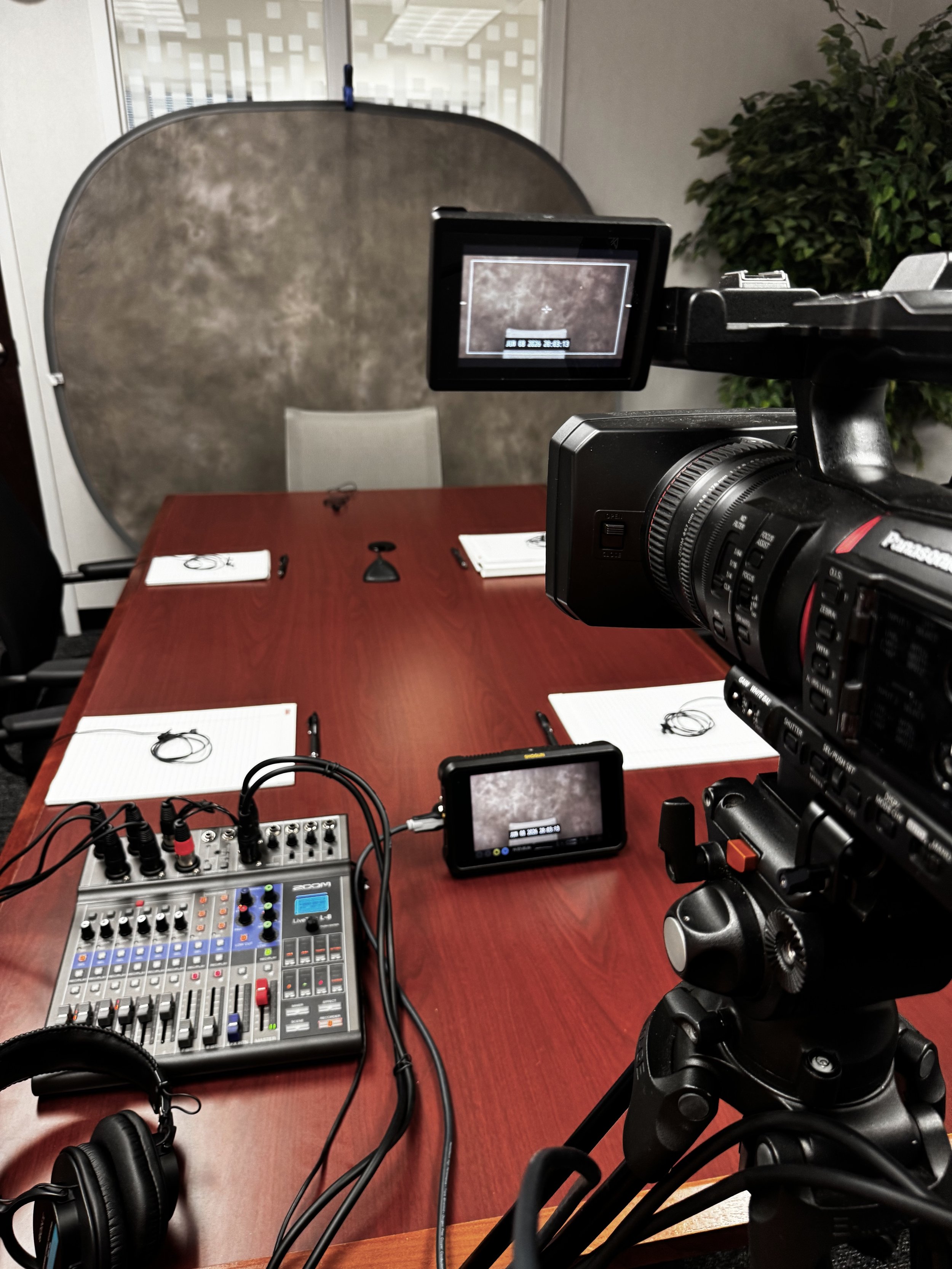 Recording setup with a professional video camera aimed at a conference room table, which has notepads, pens, and headphones, with a background of office window and green plant.