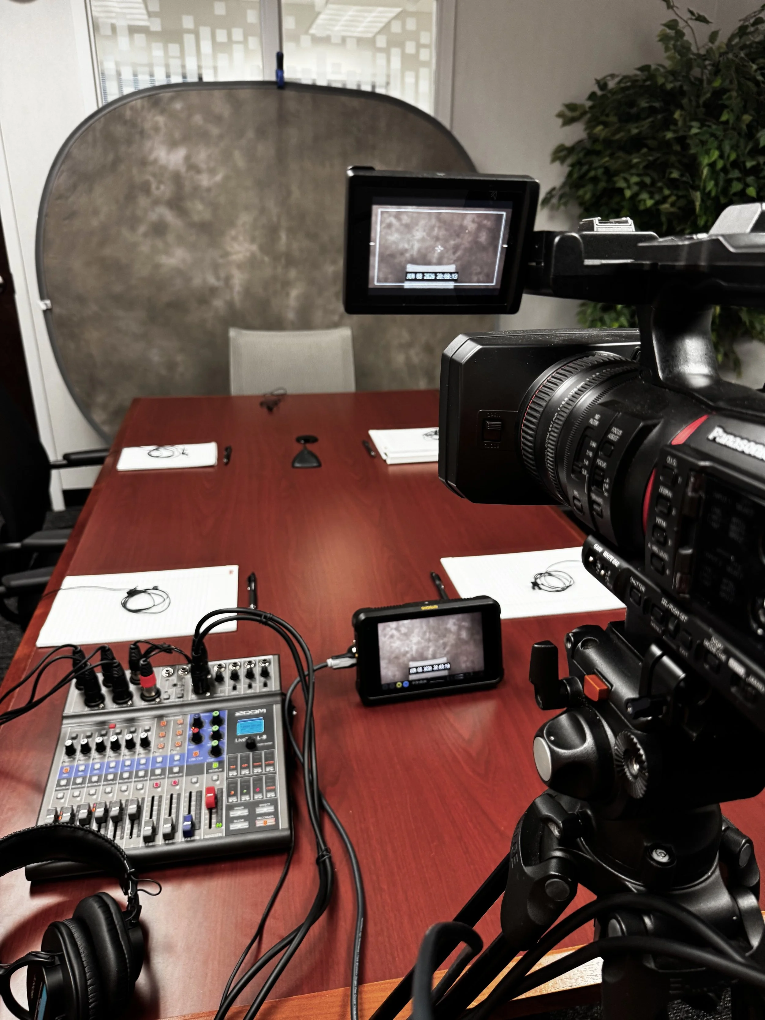 Recording setup with a professional video camera aimed at a conference room table, which has notepads, pens, and headphones, with a background of office window and green plant.