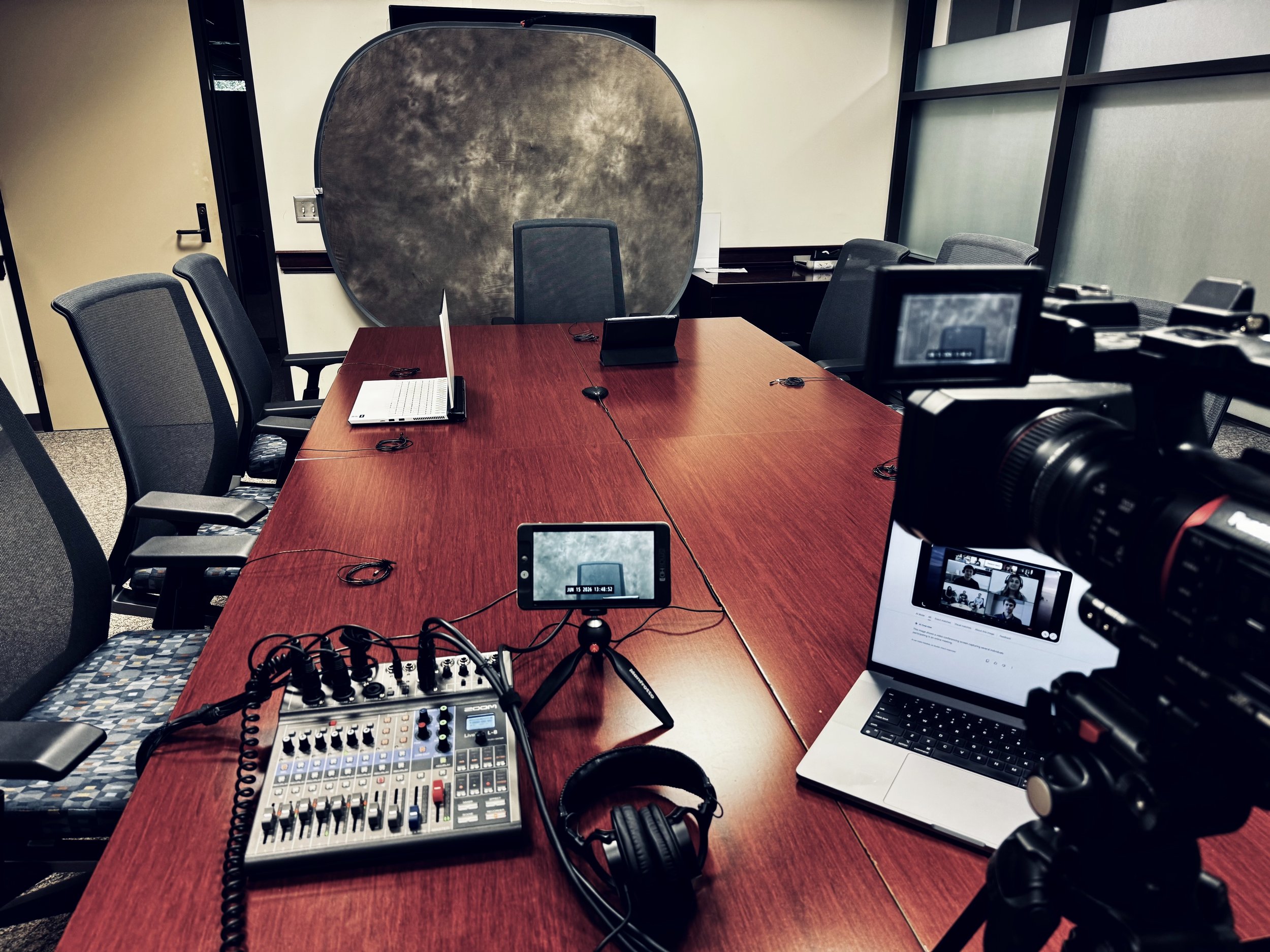 A conference room setup for a virtual meeting or recording with multiple chairs around a large wooden table, several laptops, a camera, a microphone, a mixer, headphones, and a large window with frosted glass.